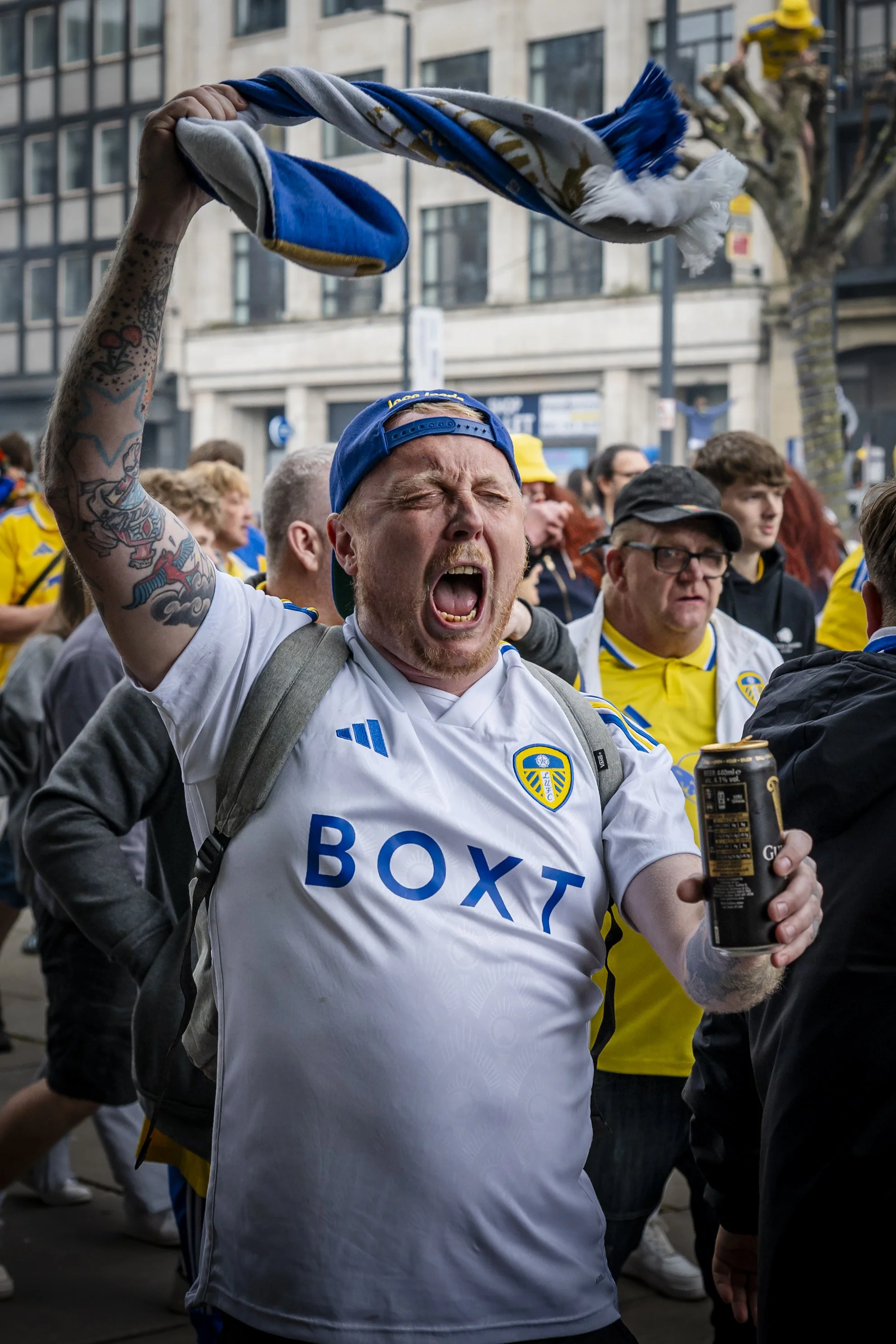 A man wearing a Leeds United football jersey and a backwards blue cap is celebrating, holding a scarf in the air and shouting passionately among a crowd during a public event or rally in an urban setting.