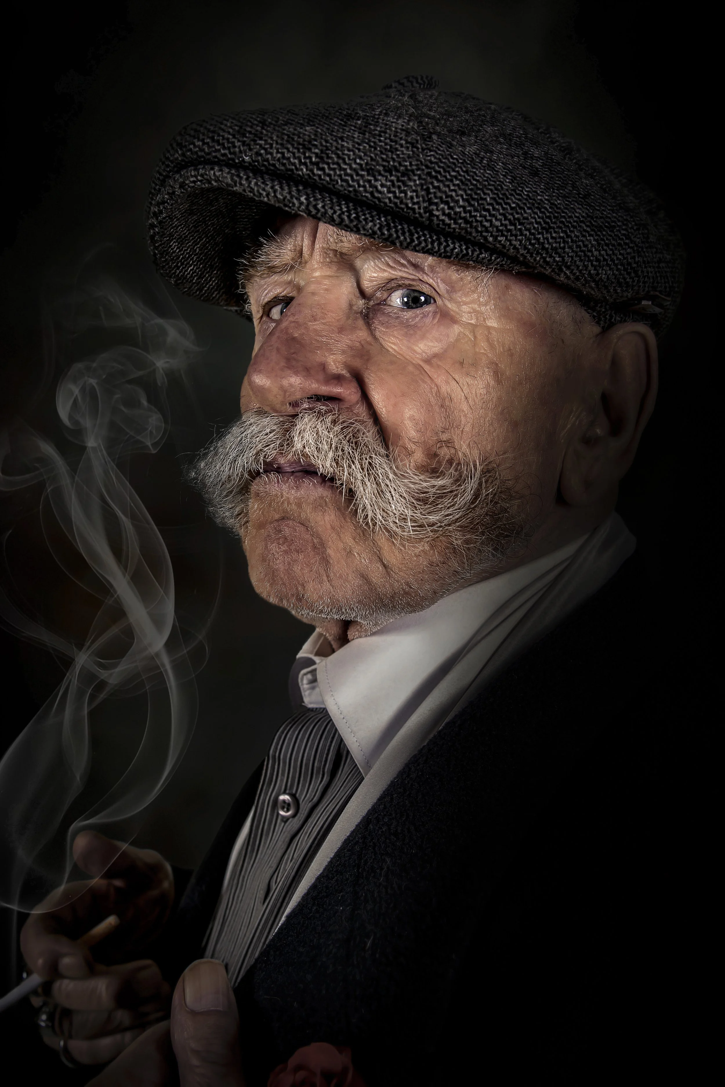 Portrait of an elderly man with a white moustache and beard, wearing a flat cap and smoking a cigarette against a dark background.
