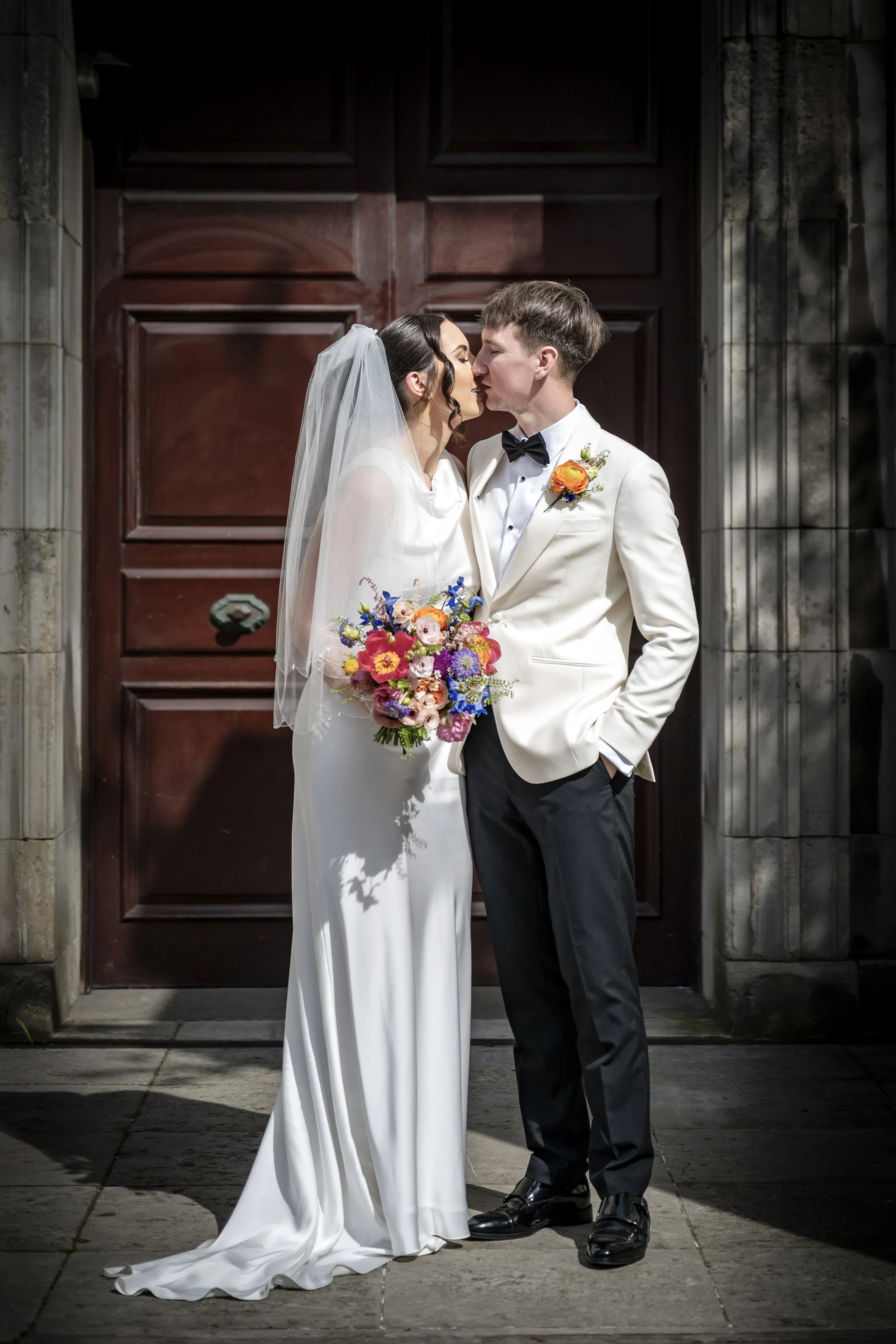 A bride and groom stand close together, about to kiss, outside a building with dark wooden doors. The bride wears a white wedding gown with a veil and holds a colorful bouquet. The groom is dressed in a white tuxedo jacket with a black bow tie and bl