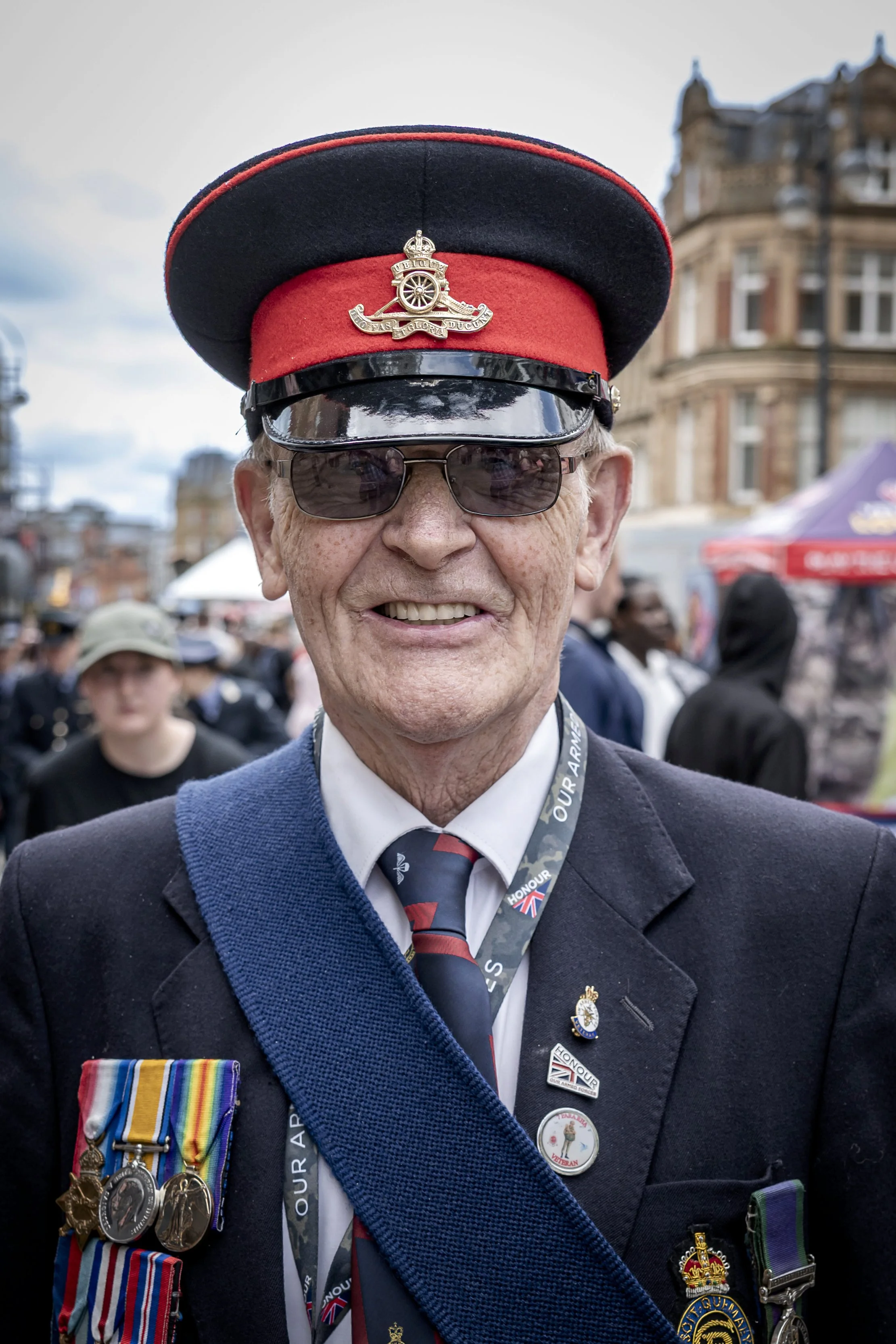 An elderly man in military attire and a hat with badges, medals, and ribbons, smiling at an outdoor event with a crowd and historic buildings in the background.