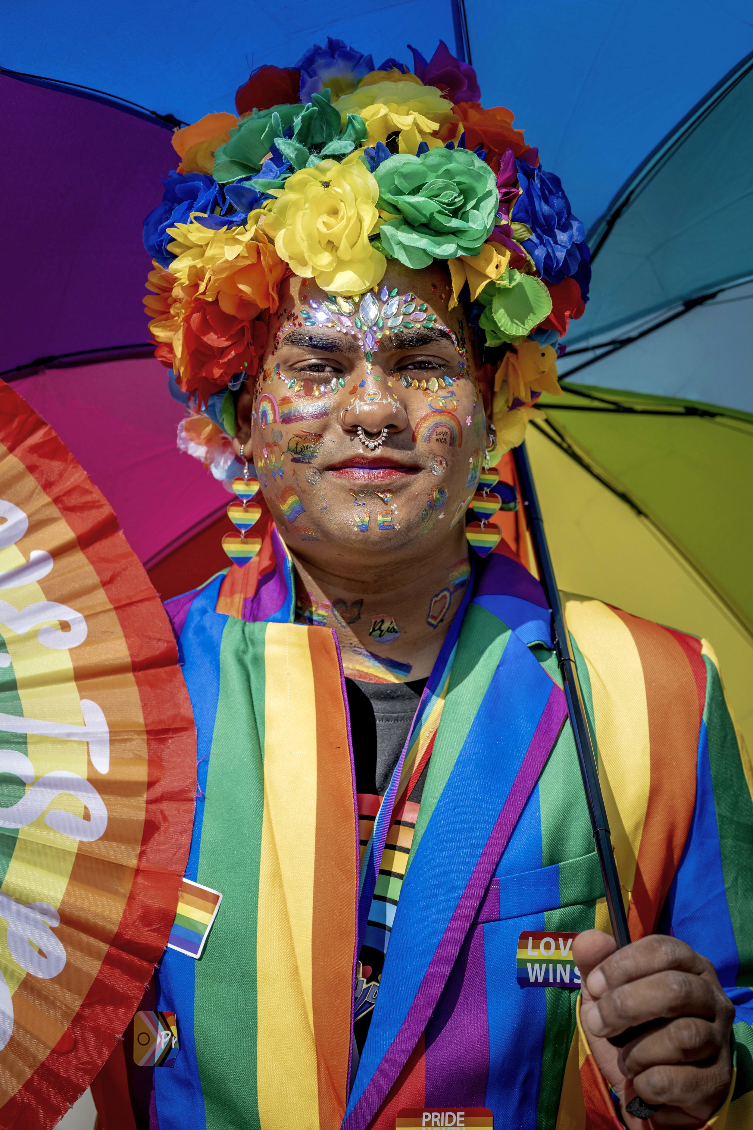 Person dressed in colorful rainbow costume with a large flower crown and rainbow face makeup, holding a rainbow umbrella. The person has rainbow-themed face and body stickers and is wearing rainbow-themed earrings and a rainbow-colored vest. The back