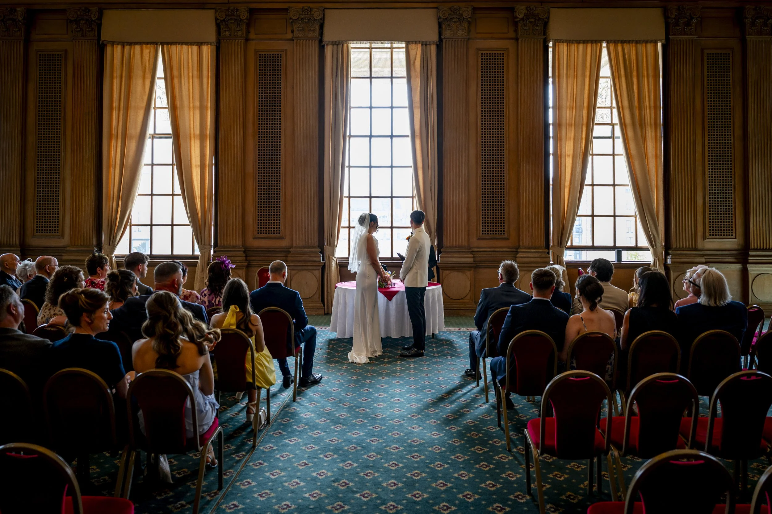 A wedding ceremony with a bride and groom standing before a judge or officiant in a grand, wood-paneled room with tall windows and gold curtains. Guests are seated, watching the couple.