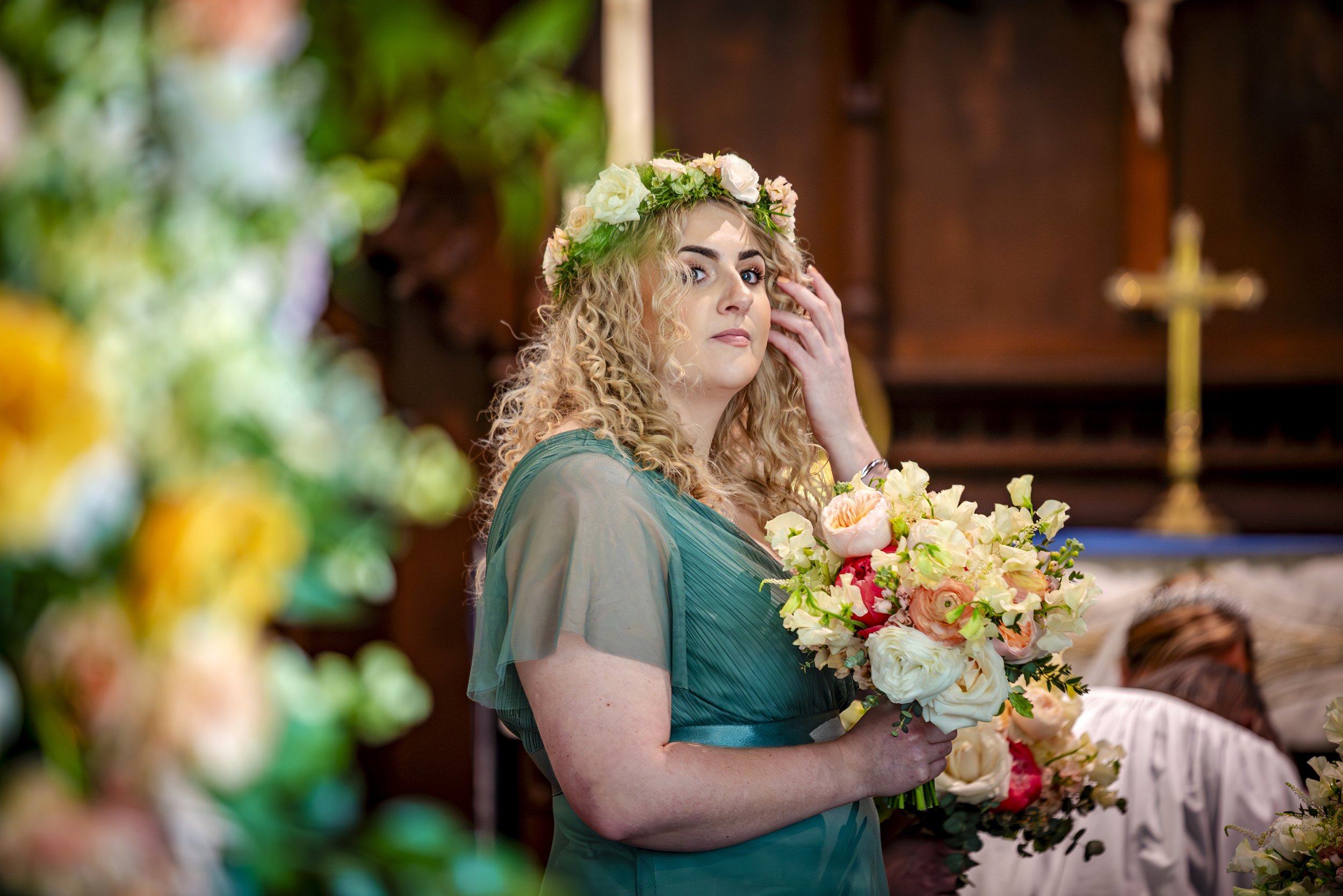 A woman with curly blonde hair in a green dress is holding a large bouquet of flowers and touching her face in a church setting, wearing a floral crown.