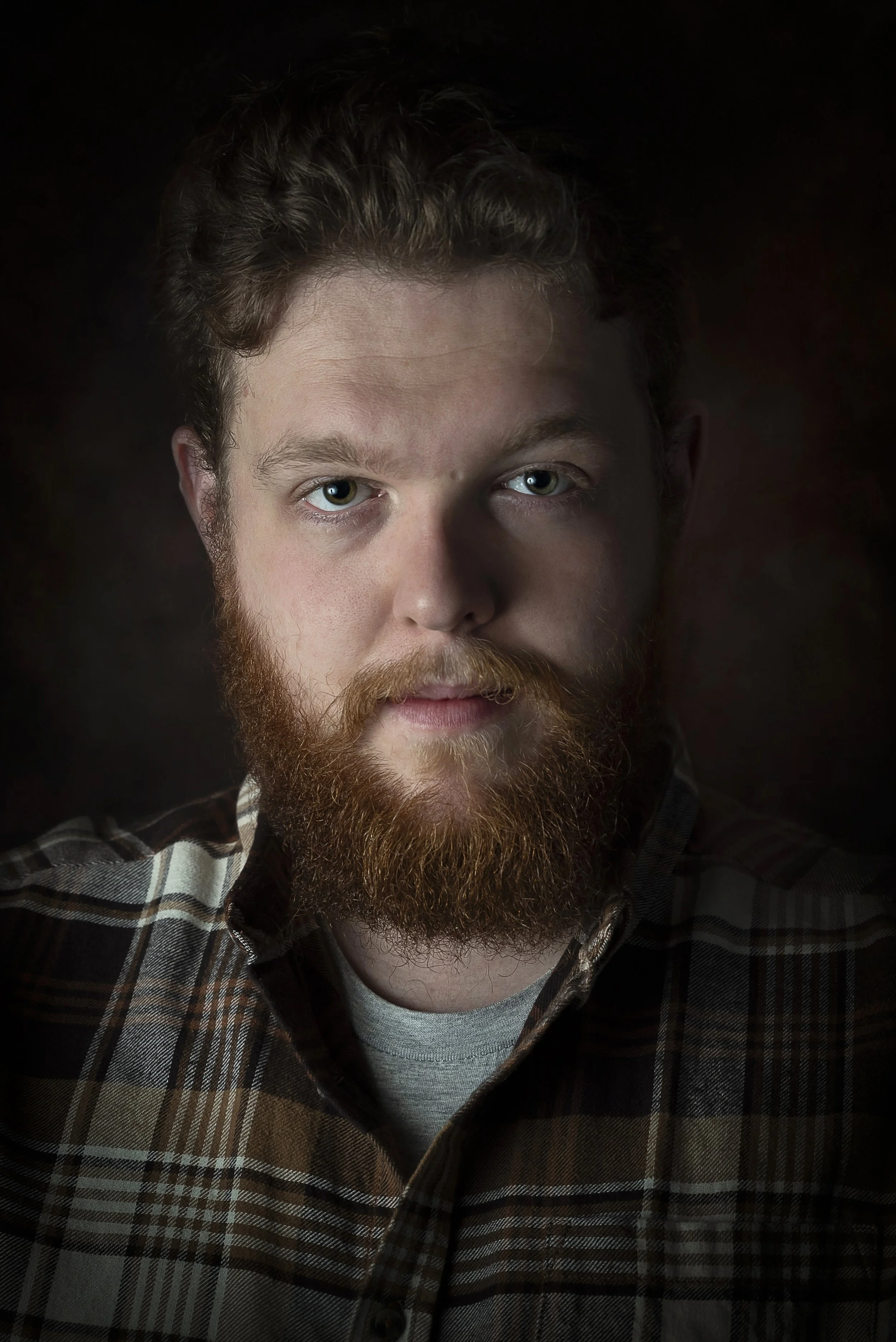 A man with red hair and a beard wearing a plaid shirt, looking directly at the camera, dark background.