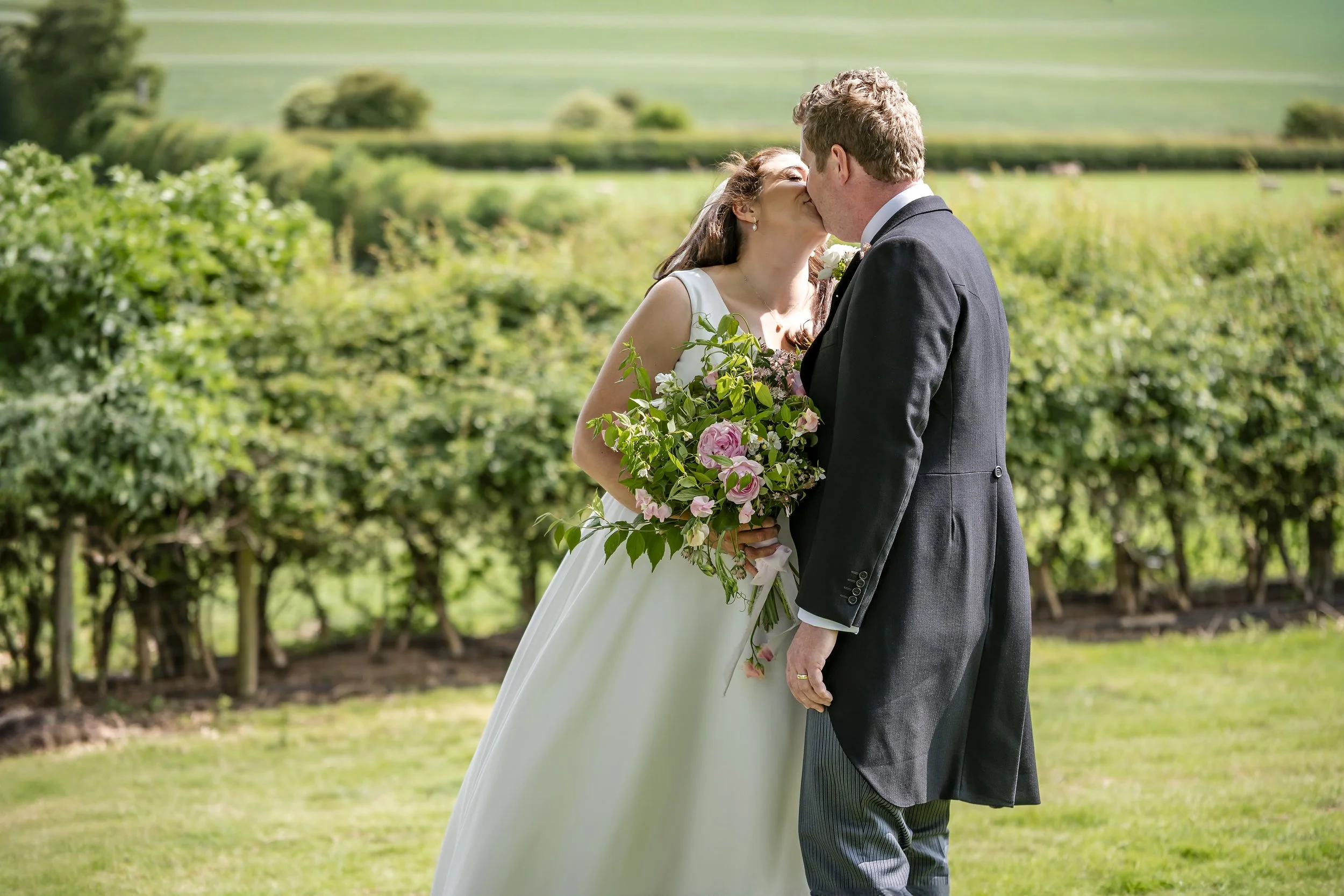 A bride and groom sharing a kiss outdoors in a garden, with the bride holding a bouquet of pink and white flowers.