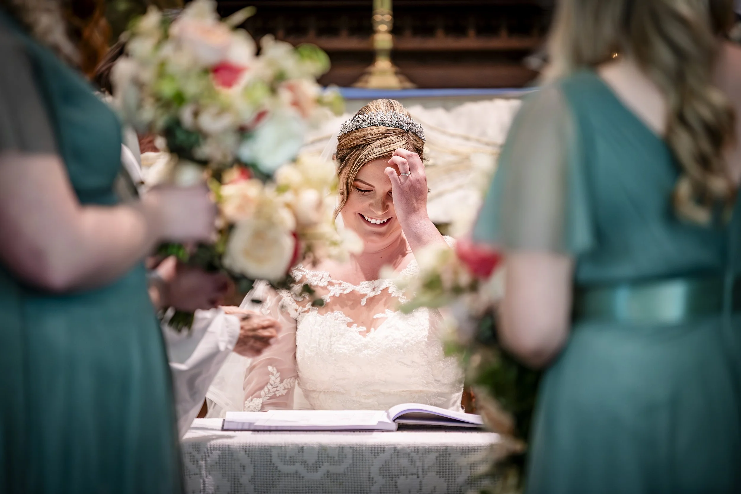 A bride in a white wedding dress and tiara smiling with her hand on her forehead during a wedding ceremony, surrounded by bridesmaids holding bouquets.