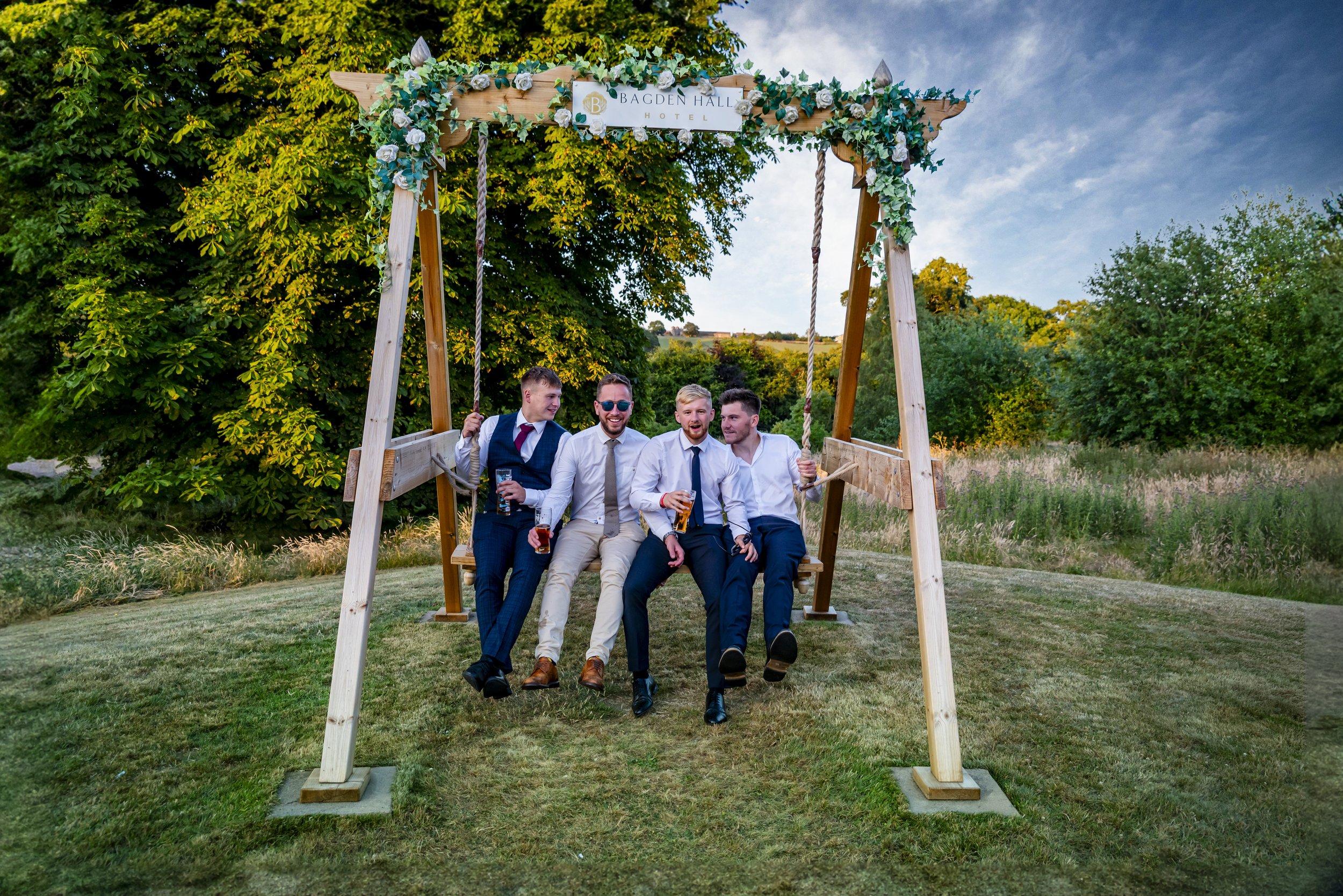 Four men in formal attire sitting on a large wooden swing at an outdoor event during sunset, holding drinks and laughing, with trees and a field in the background.