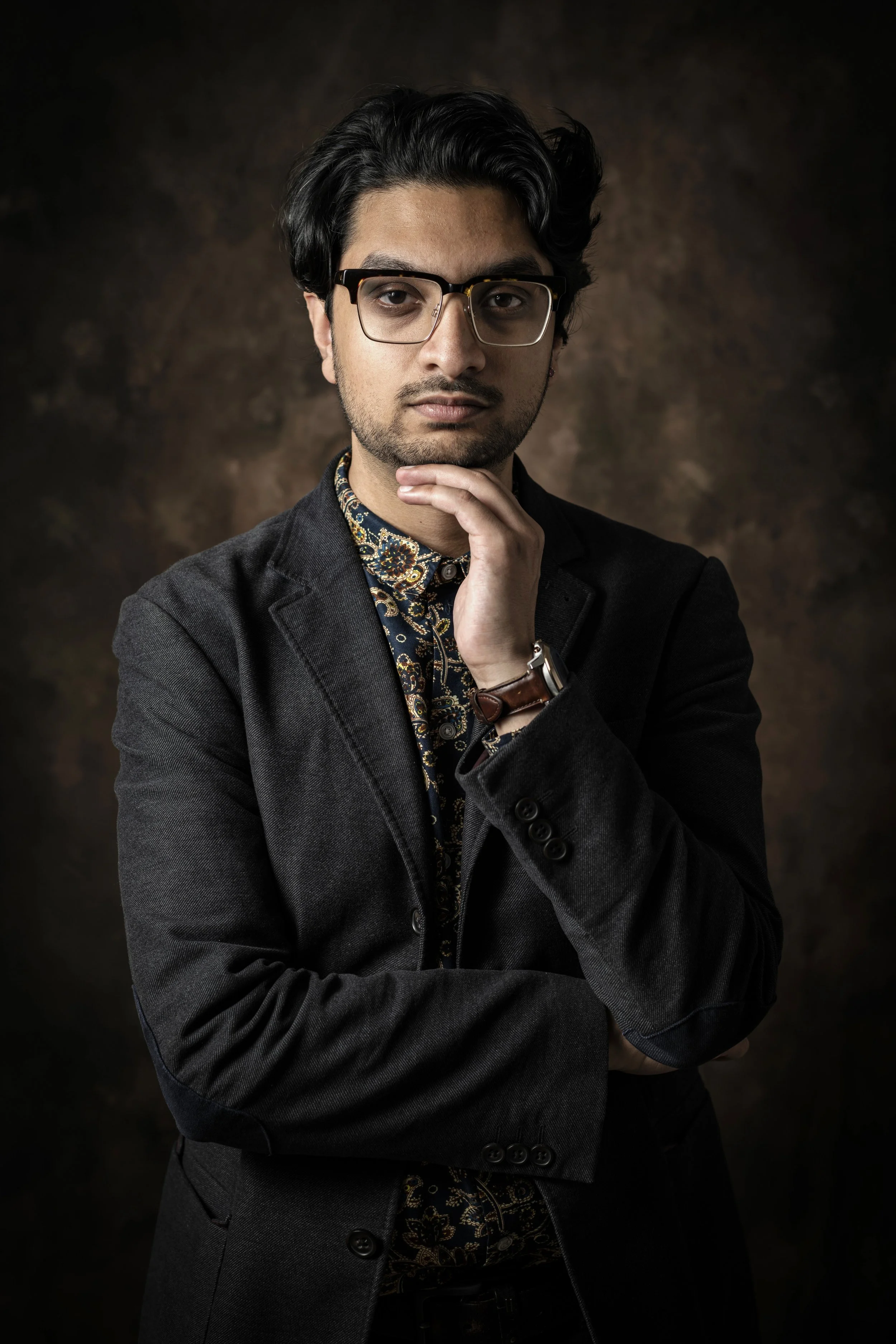 A young man with dark hair and glasses, wearing a black blazer, patterned shirt, and watch, posing against a dark textured background.