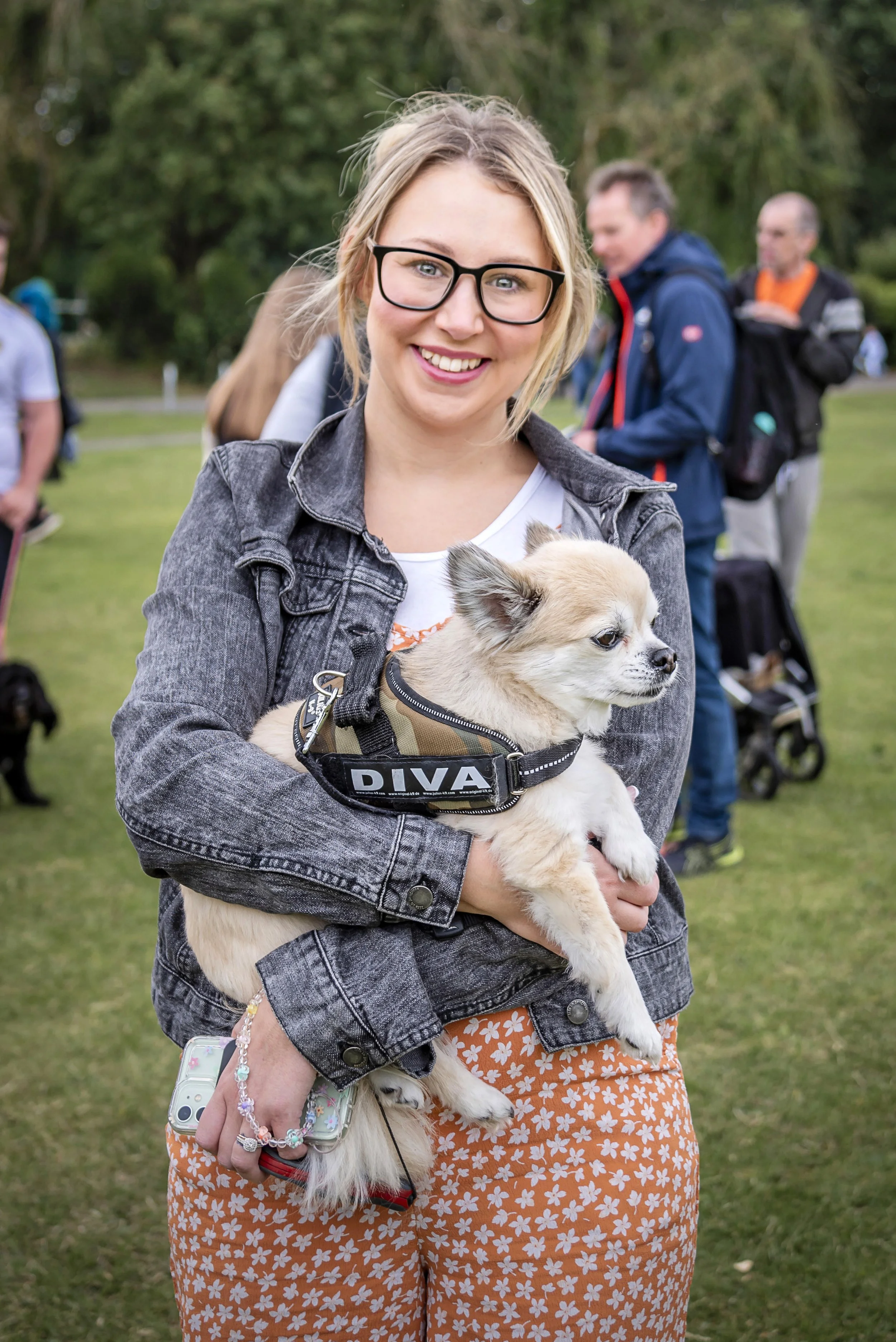 A young woman with glasses smiling while holding a small beige and white dog wearing a harness with the word 'DIVA' on it, outdoors in a park with green trees and other people in the background.