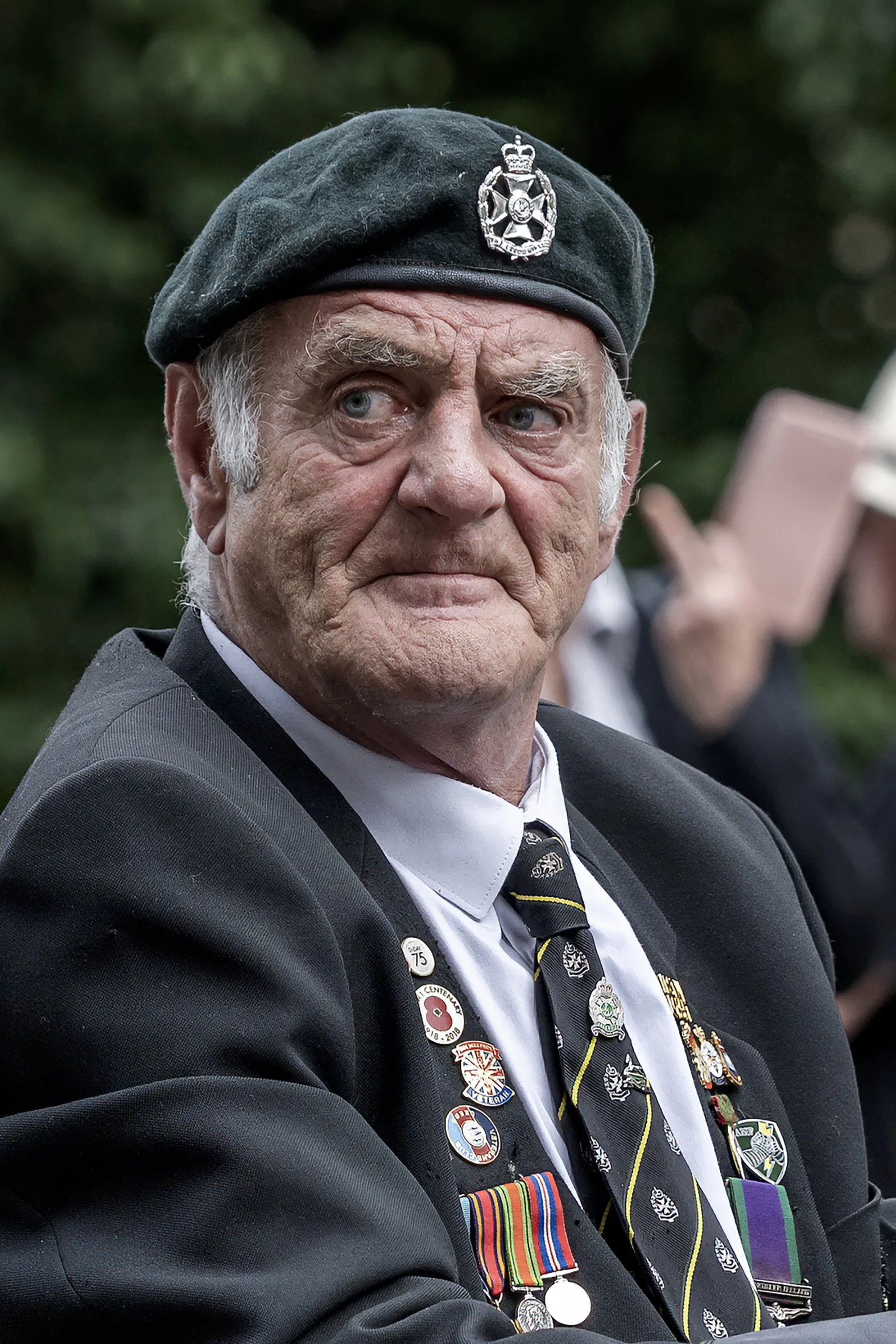 An elderly man with a serious expression wearing a grey beret with a badge, a black suit, and a white shirt. His jacket is decorated with various military medals and ribbons. The background is outdoors with green blurred foliage.