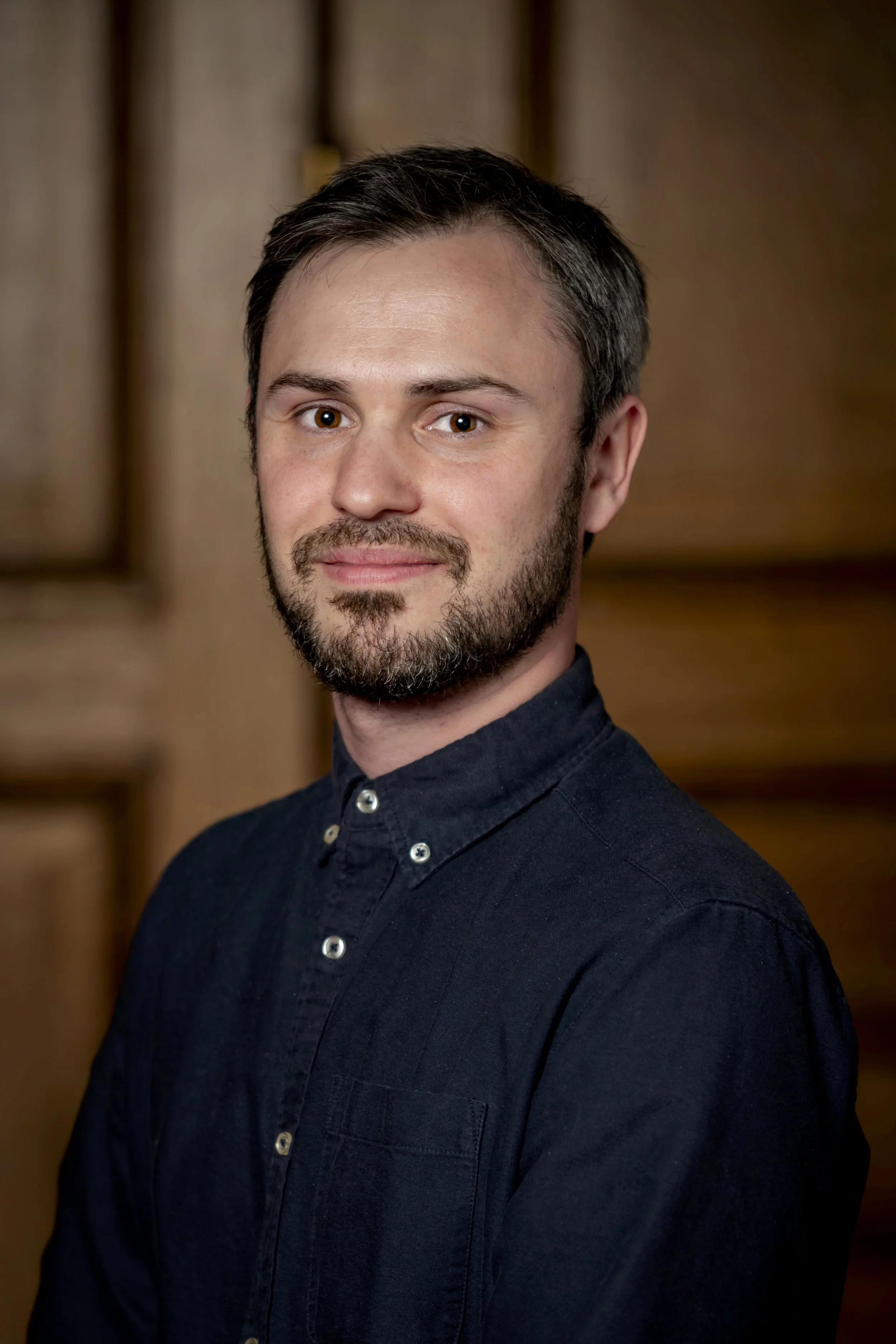 A portrait of a man with dark hair, a short beard, and wearing a dark button-up shirt, standing in front of a wooden background.