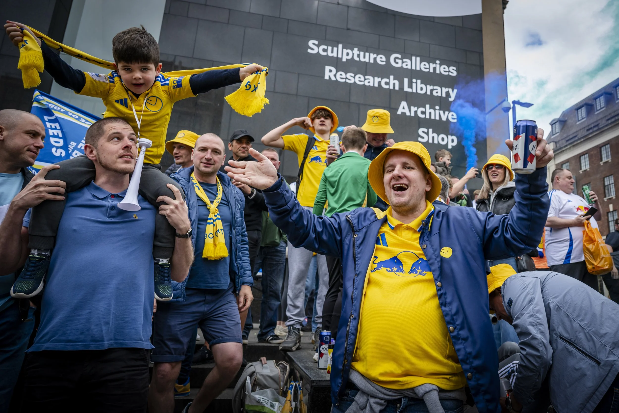 A group of fans dressed in yellow and blue clothing celebrating outside a building with a sign that reads 'Sculpture Galleries Research Library Archive Shop.' One person is sitting on someone else's shoulders, holding a scarf, while others are holdin