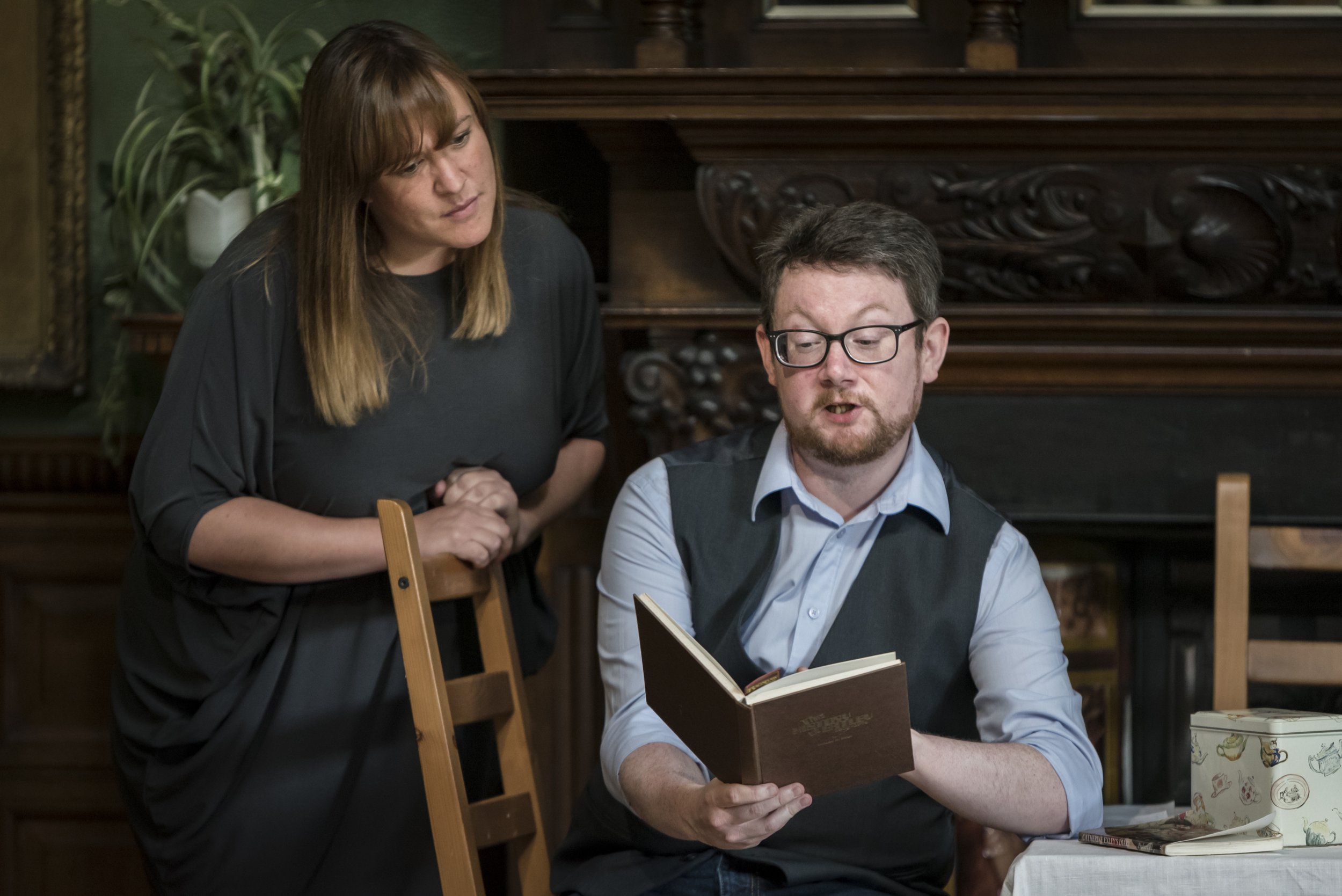 A woman and a man are looking at a menu in a restaurant. The woman is leaning over the man, who is seated and reading the menu. The background includes a dark wooden piece of furniture and a green plant.
