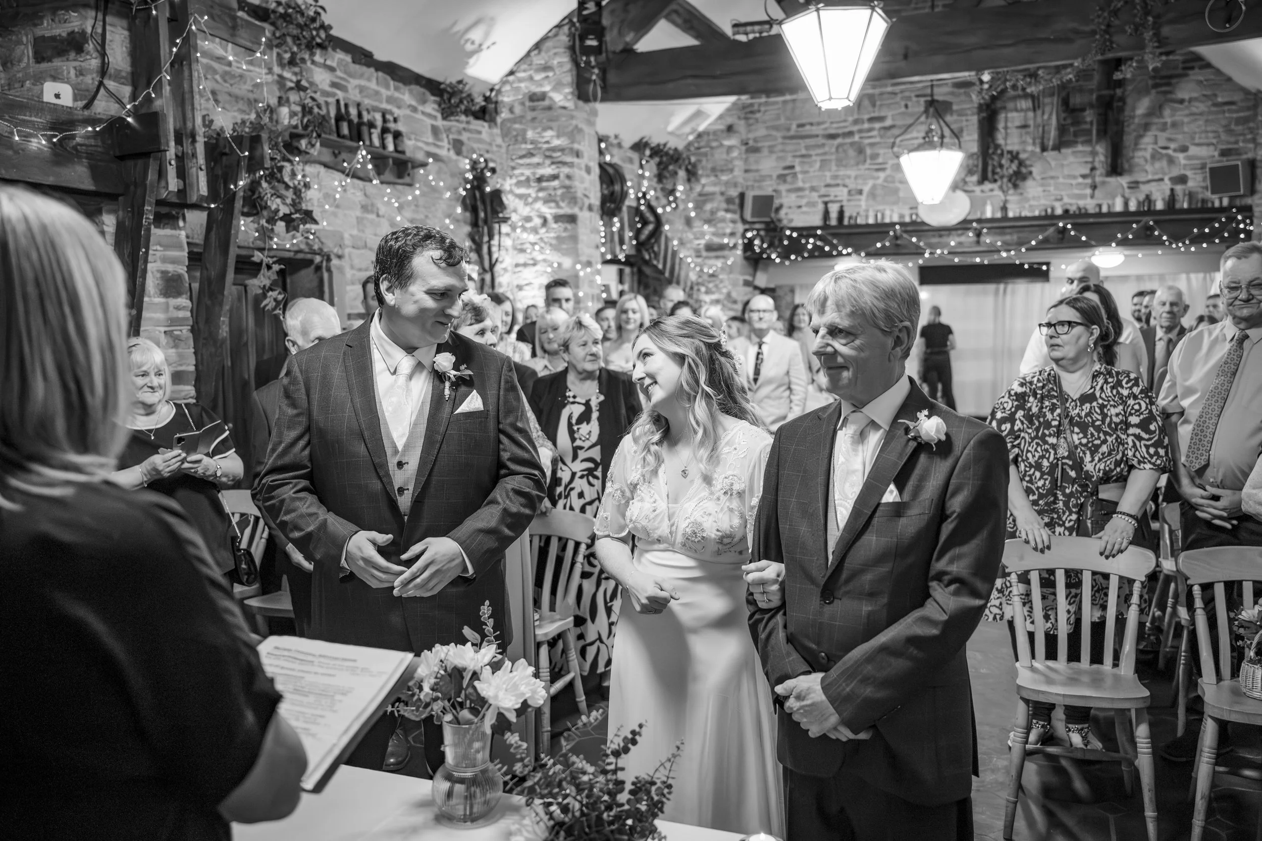 A wedding ceremony with a bride and groom standing in front of an officiant. The bride is smiling at an older man, likely her father, as he holds her hand. The groom looks at the bride. The scene is inside a rustic venue with stone walls, string ligh
