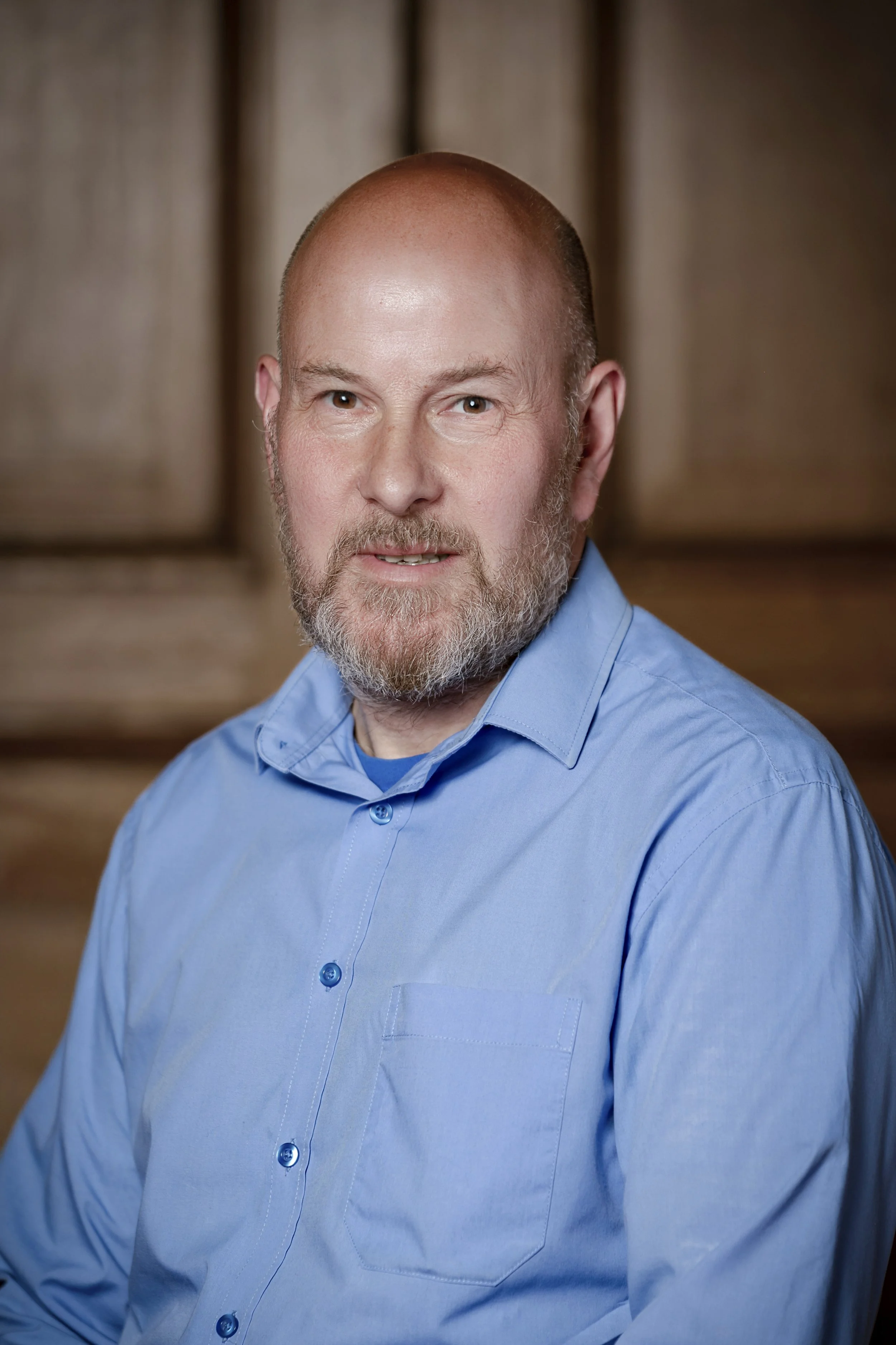 A middle-aged man with a beard and bald head wearing a blue button-up shirt, posing for a professional headshot with a wooden background.