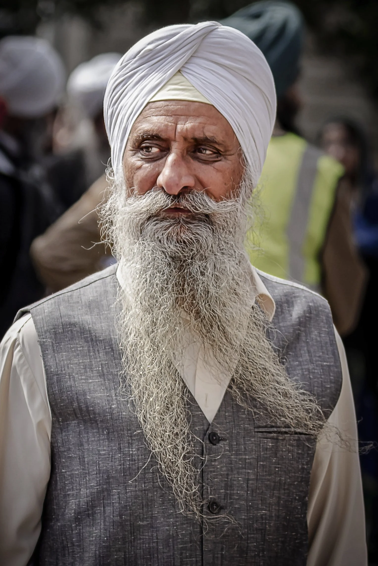 An elderly man wearing a white turban and traditional Indian attire with a long beard stands among a group of people in the background.