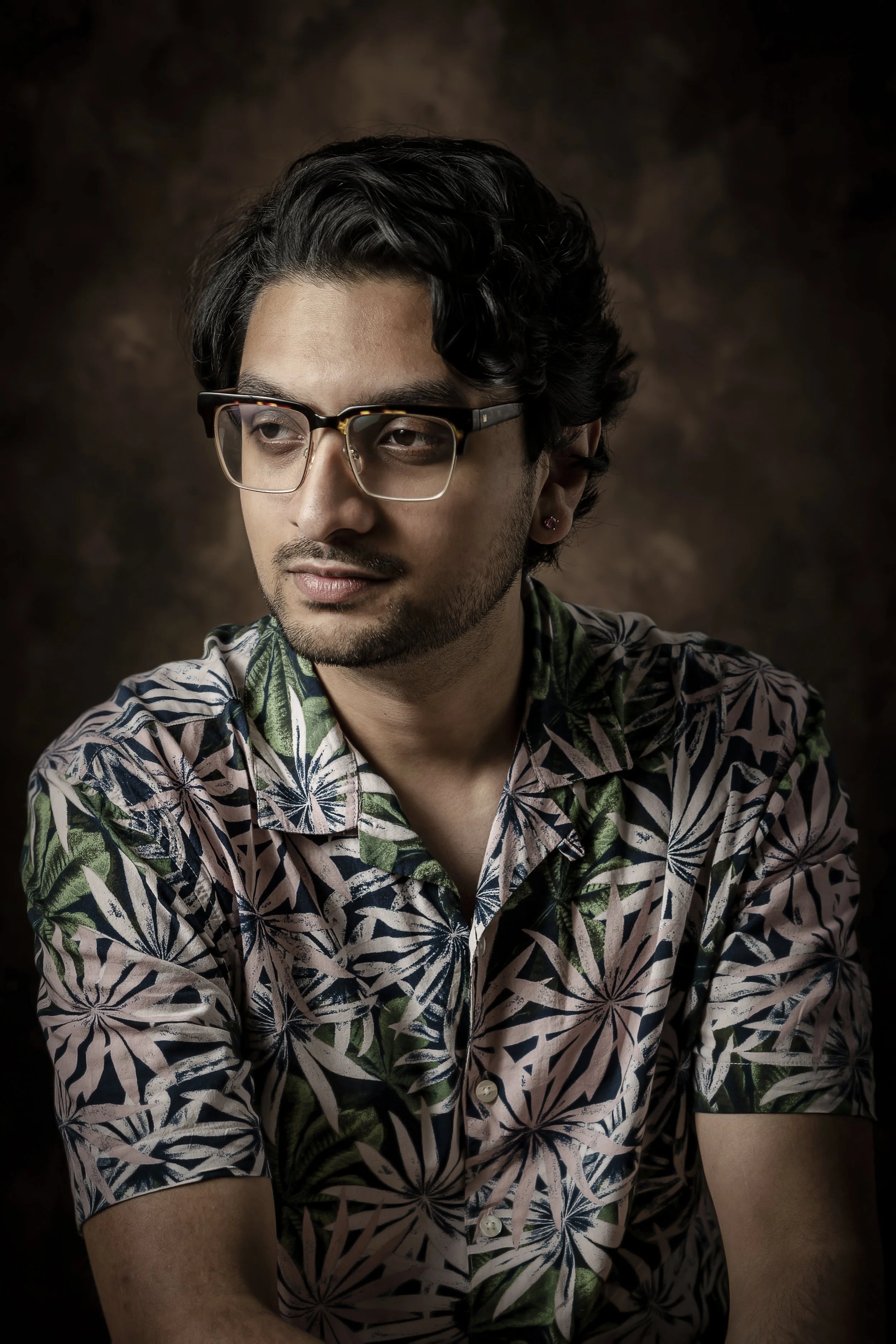 A young man with wavy black hair, glasses, and a small earring in one ear, wearing a colorful floral shirt, looking to the side with a neutral expression, against a dark brown background.