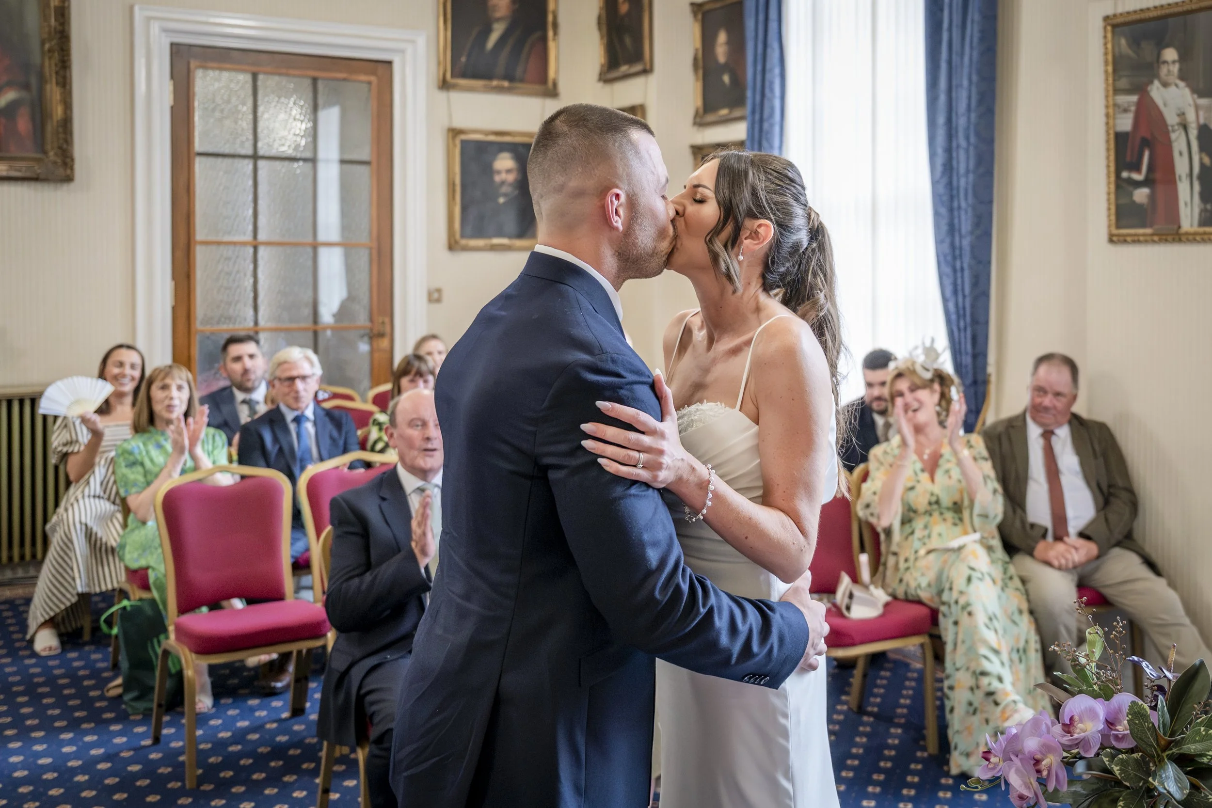 A newlywed couple sharing a kiss during their wedding ceremony, surrounded by seated guests, in a room decorated with portraits and blue curtains.