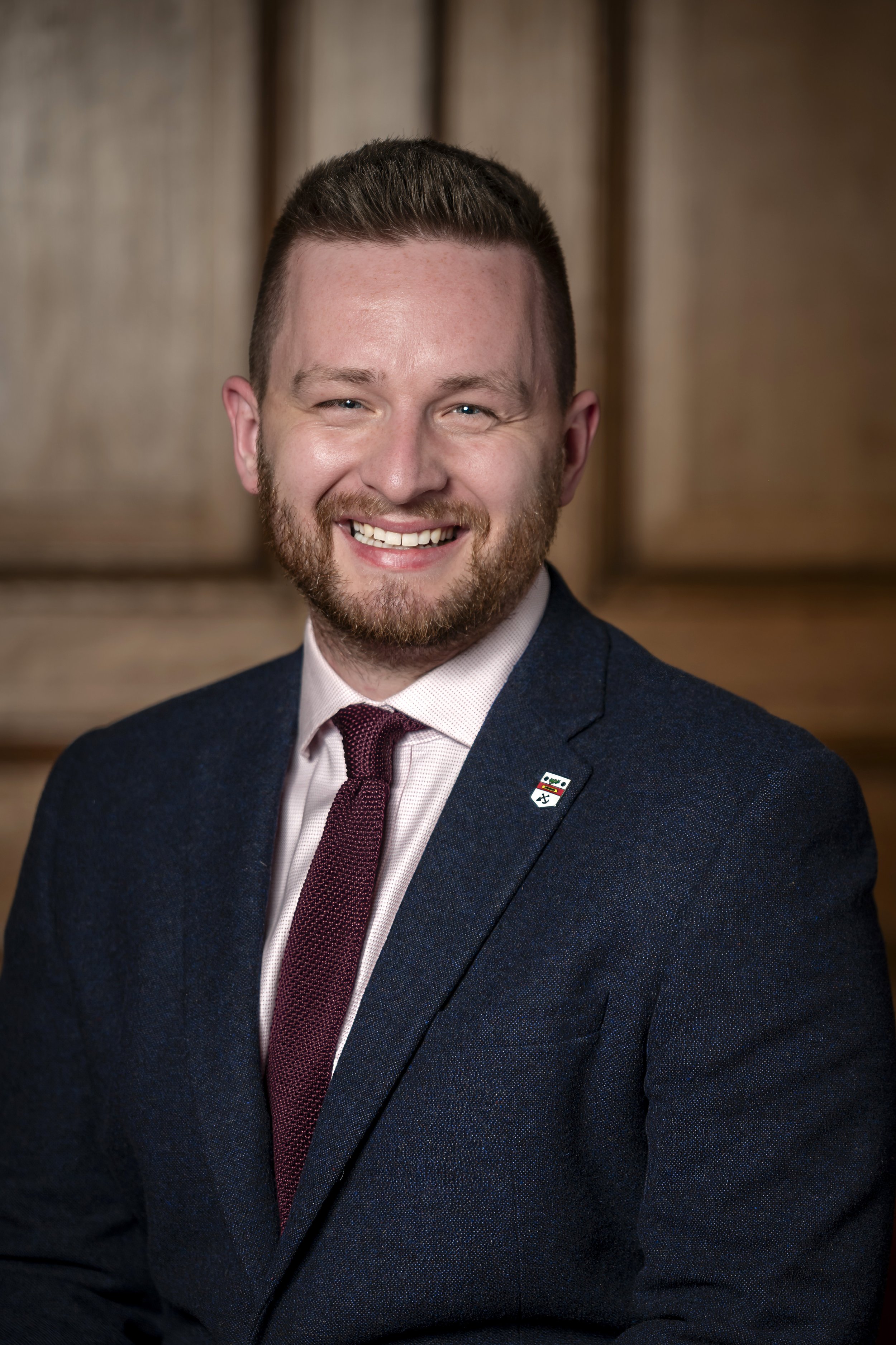 Young man with short hair and beard, wearing a navy suit, light pink shirt, maroon tie, and a small lapel pin, smiling against a wooden background.