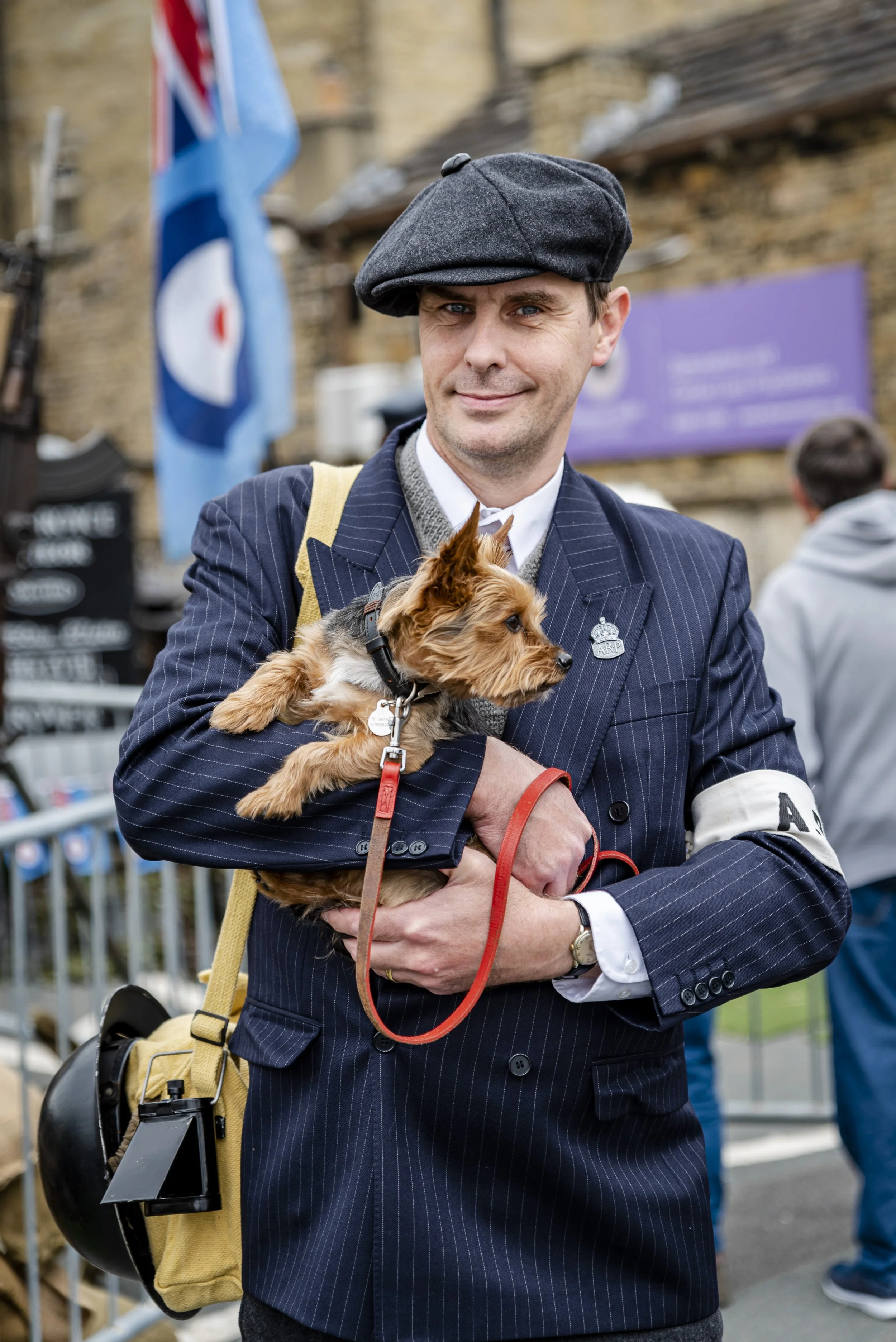 Man in a vintage navy blue pinstripe suit holding a small dog, wearing a flat cap and a lapel pin, with a yellow bag and a helmet hanging on his side, outdoors with a flag in the background.