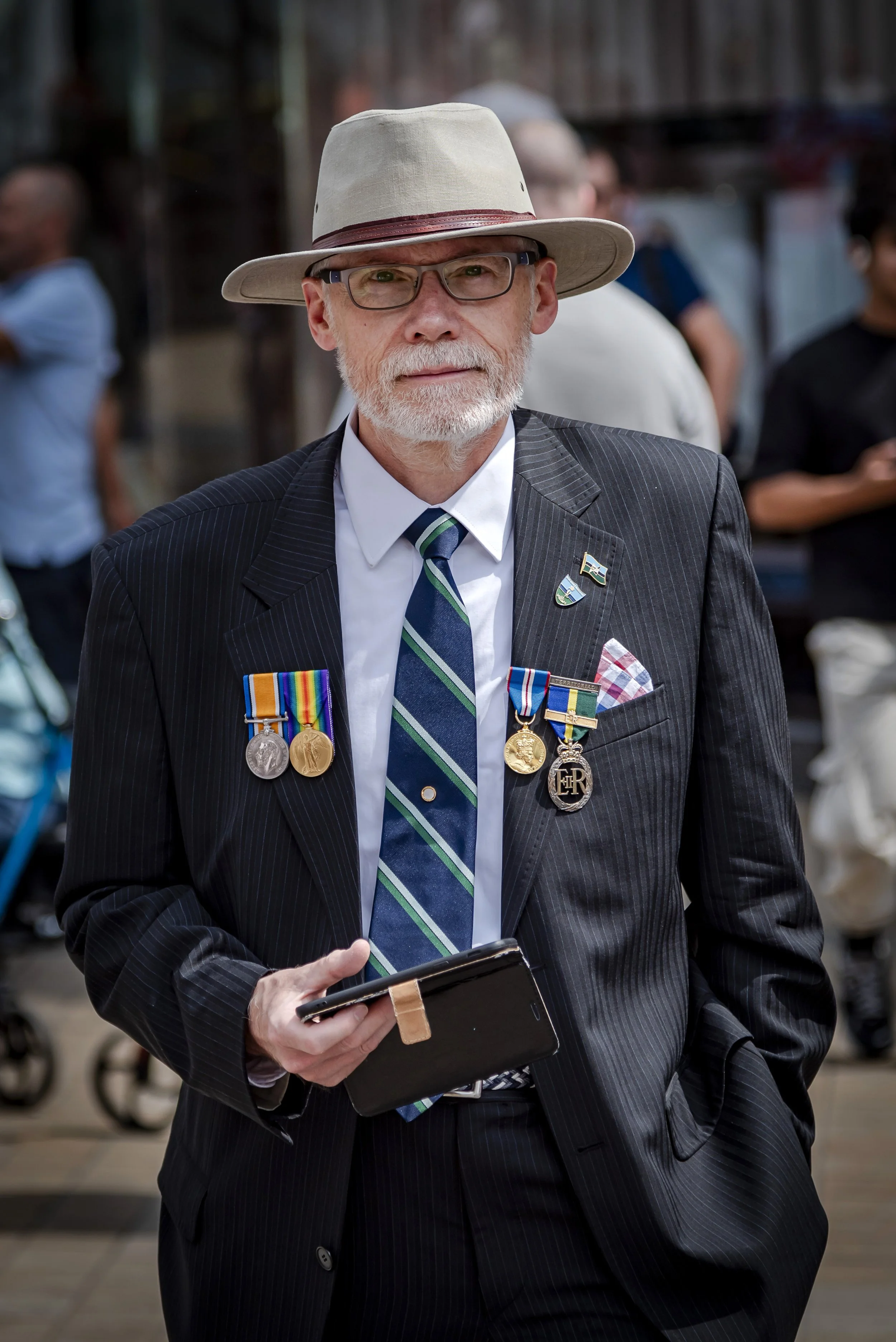 A distinguished older man wearing a suit with medals, a tie, glasses, and a wide-brimmed hat, standing outdoors in a crowd.