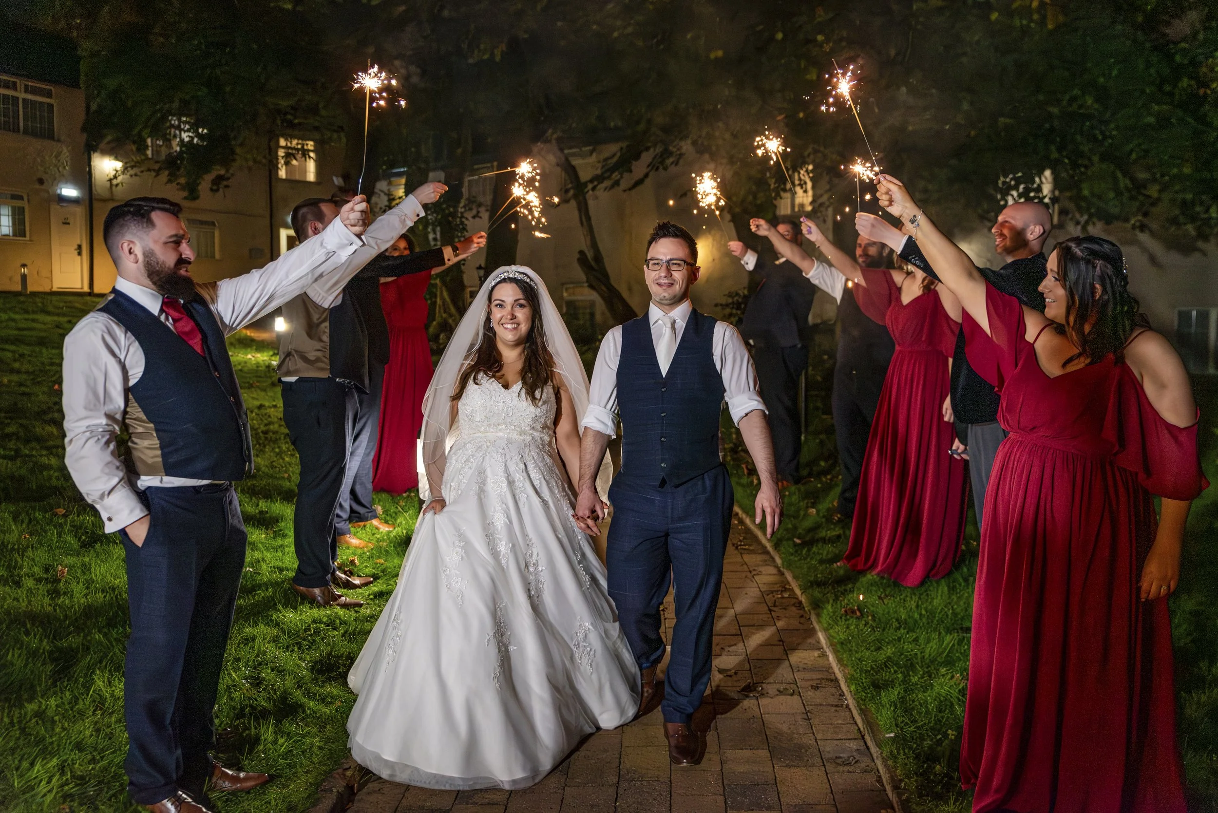 A bride and groom holding hands walking on a sidewalk at night. They are surrounded by friends holding sparklers, celebrating a wedding. The bride is wearing a white wedding dress with a veil, and the groom is dressed in a vest with white shirt. Frie