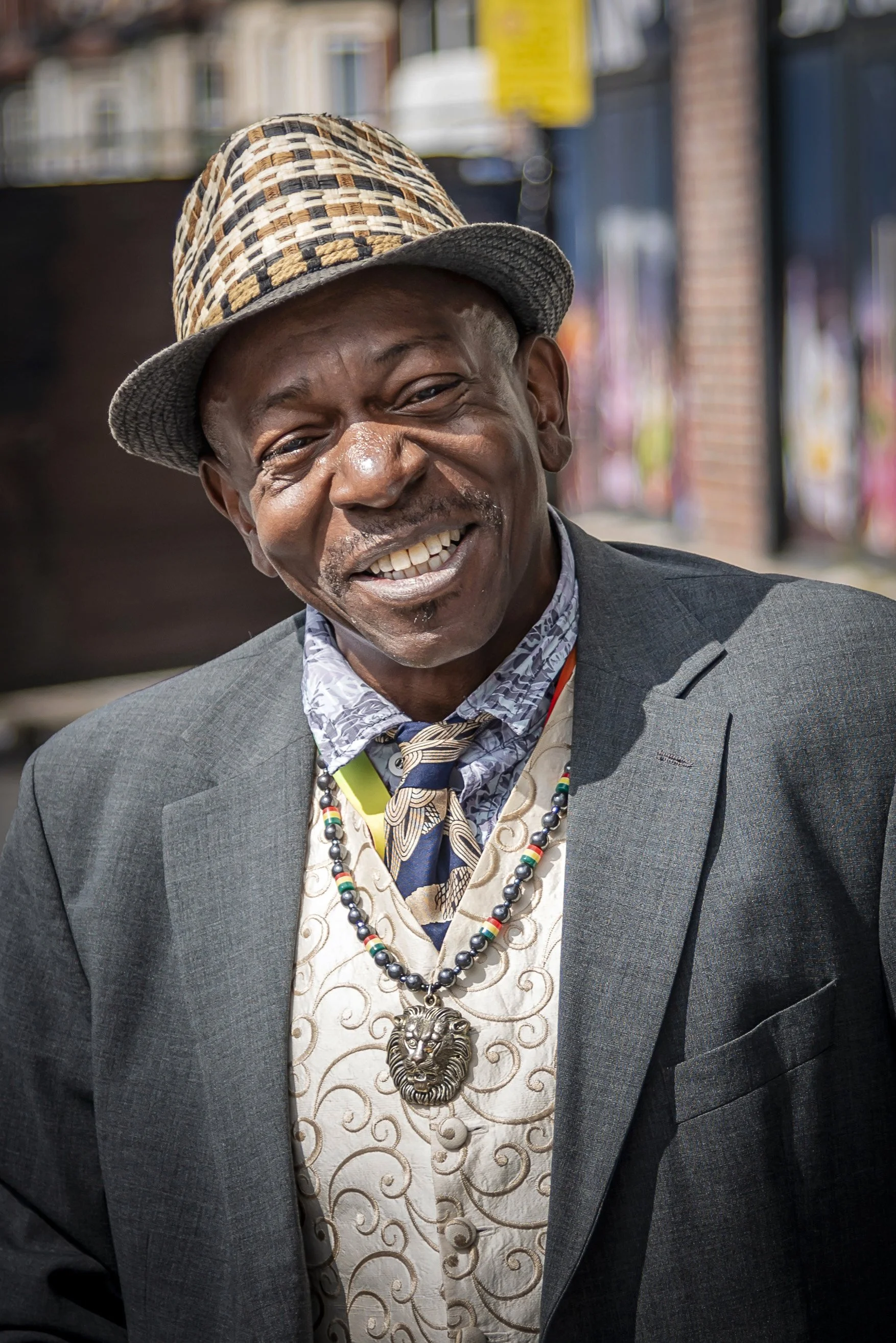 A smiling man wearing a suit, patterned shirt, colorful necklace with a lion pendant, and a checkered fedora hat, standing outdoors with a blurred background.