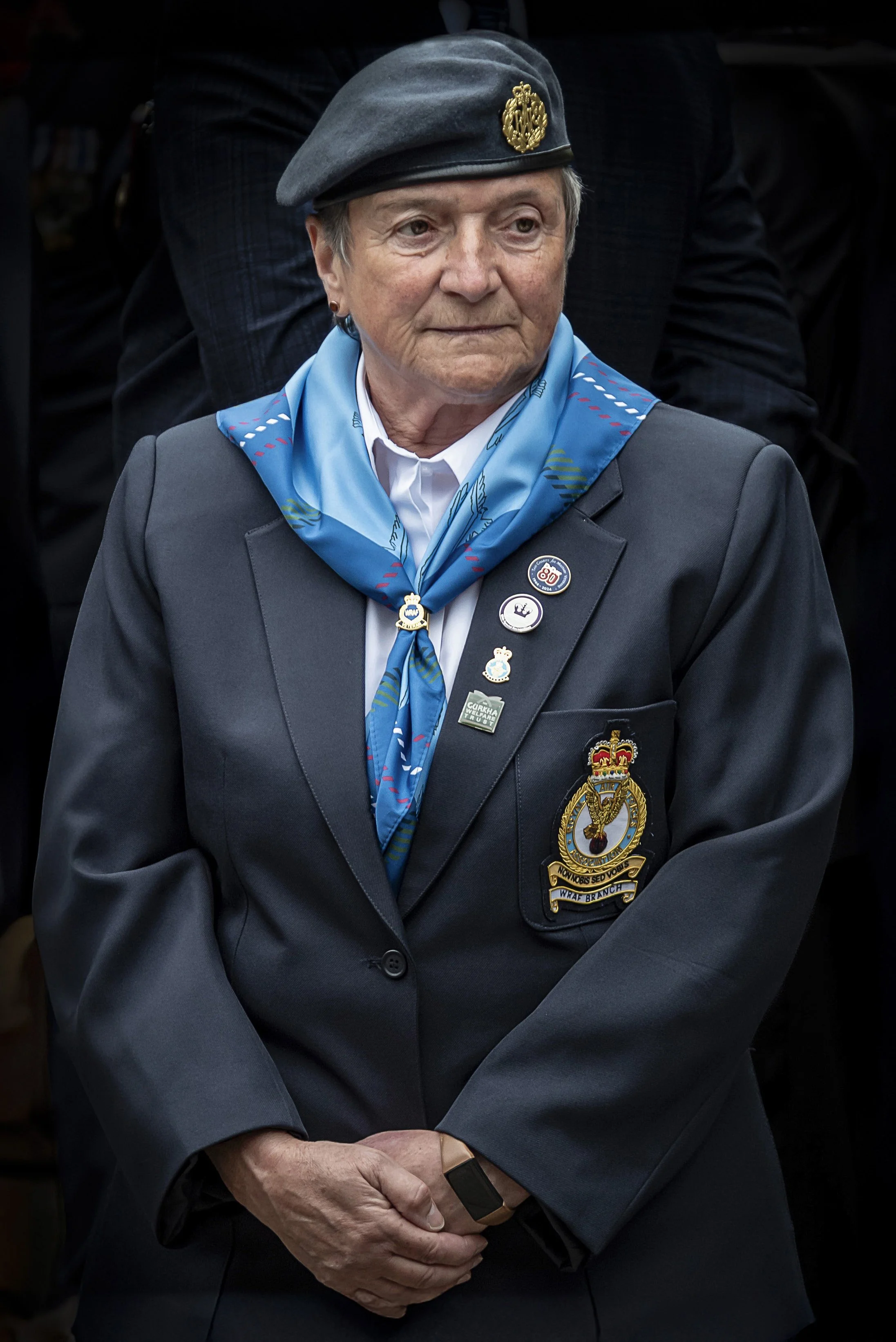 Older woman wearing a gray beret, blue neck scarf, dark blazer with badges, and a coat of arms patch, standing with hands clasped, attending a formal event.