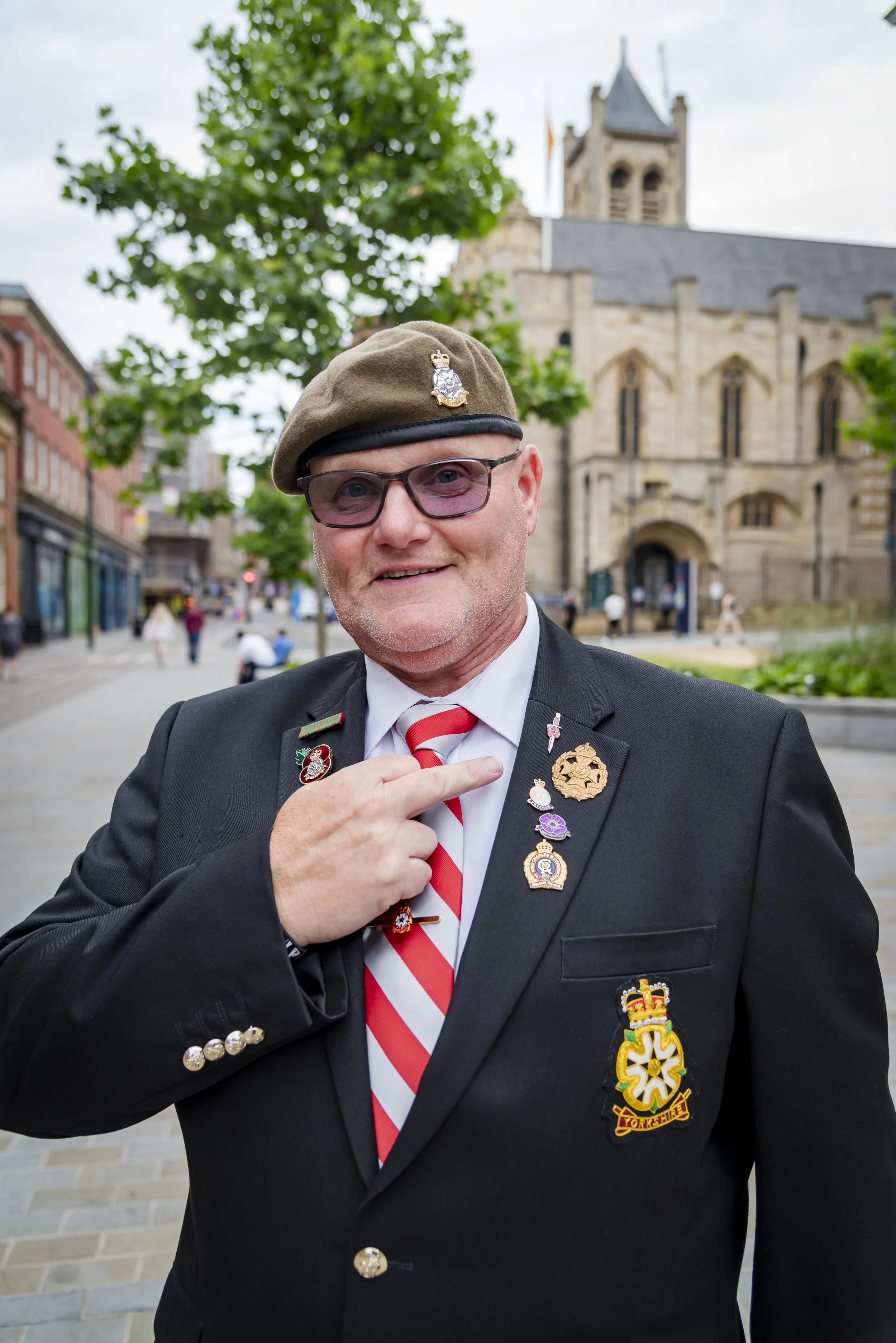 A man wearing a black suit with several medals pinned to his chest, a white shirt with a red and white striped tie, glasses, and a brown beret with a badge, standing on a city street in front of a church with a stone facade and a bell tower, with tre