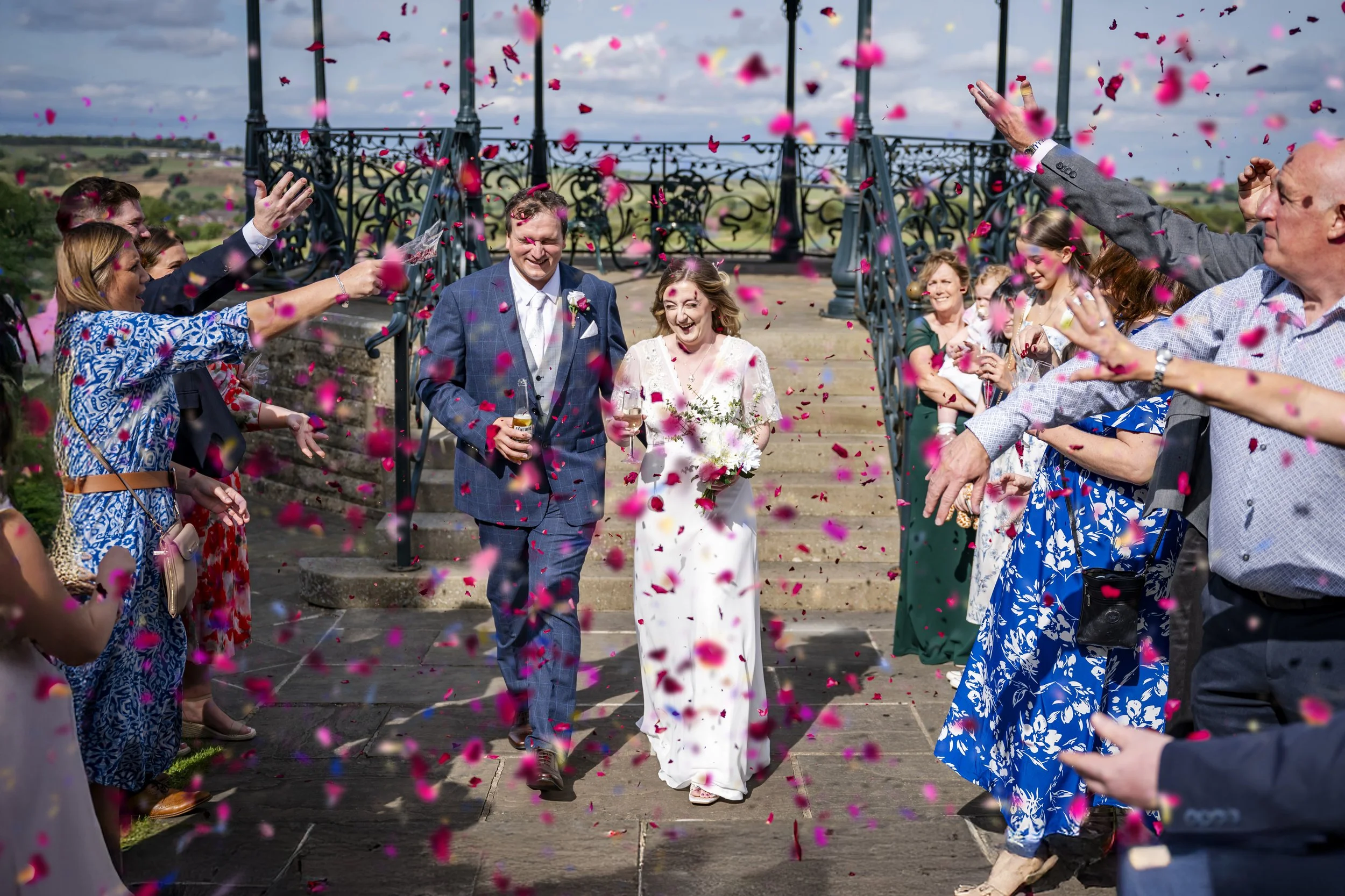 A bride and groom walking through a shower of pink and red flower petals after their wedding ceremony, surrounded by smiling guests.