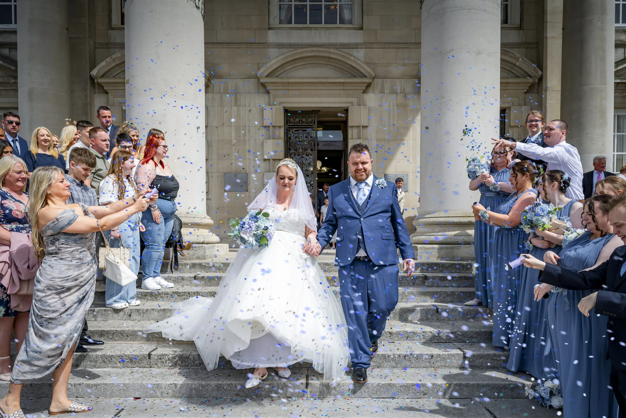 A newly married couple walking down the steps of a building after a wedding ceremony, surrounded by guests throwing blue and white confetti.