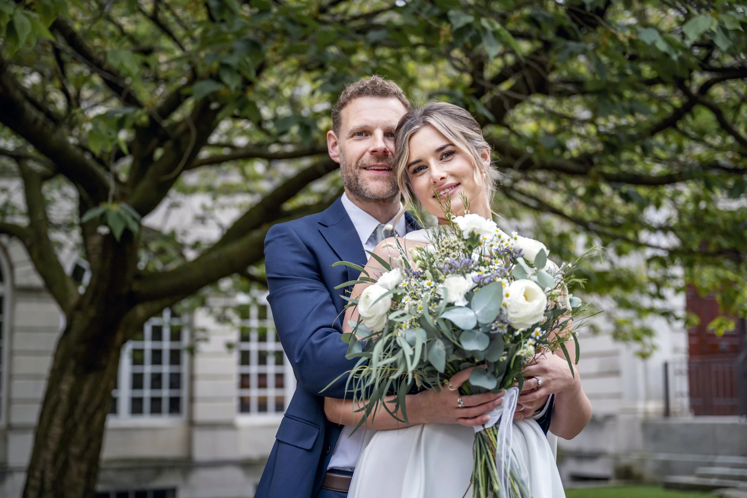 A newlywed couple embracing outdoors, with the groom in a blue suit holding a bouquet of white and purple flowers, standing under a large tree with green leaves.