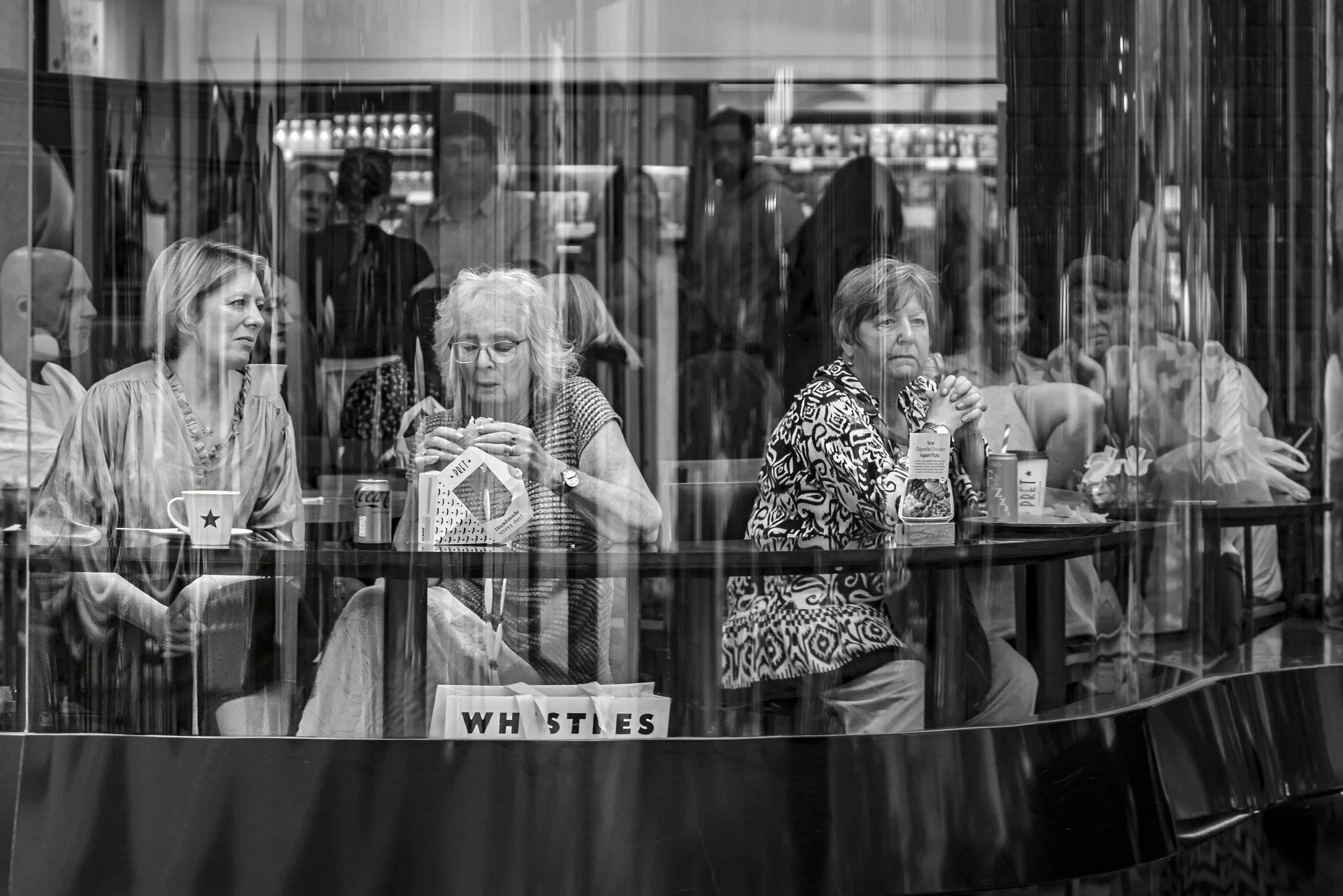 People sitting inside of a cafe, looking through a glass window. Three women are seated at a table, one has her hands clasped, another is eating, and the third is talking. In the background, other patrons and baristas are visible. The scene is in bla