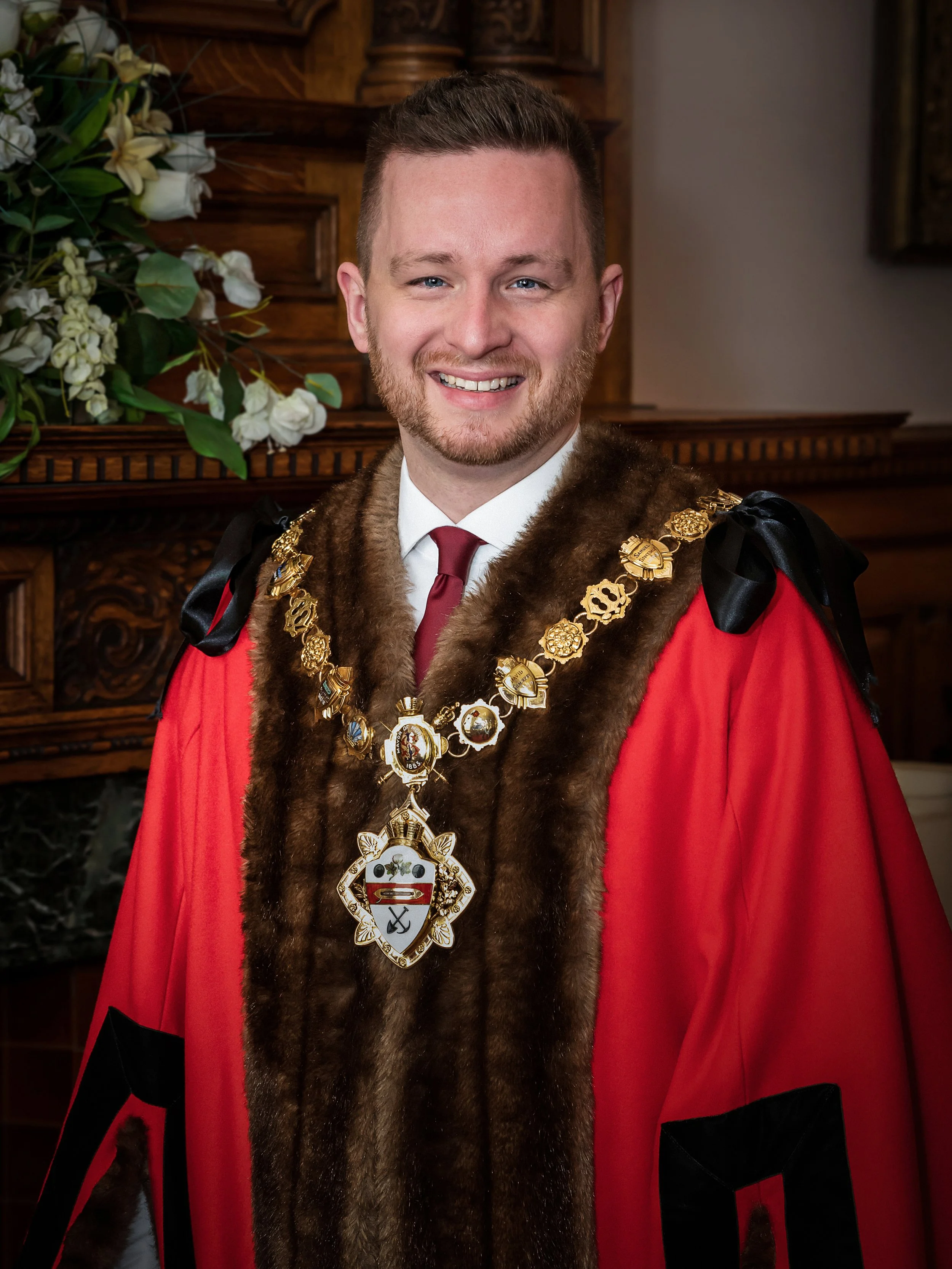 A man wearing formal robes with ornate gold chains and a large emblem, standing indoors with wood paneling and floral decorations in the background.