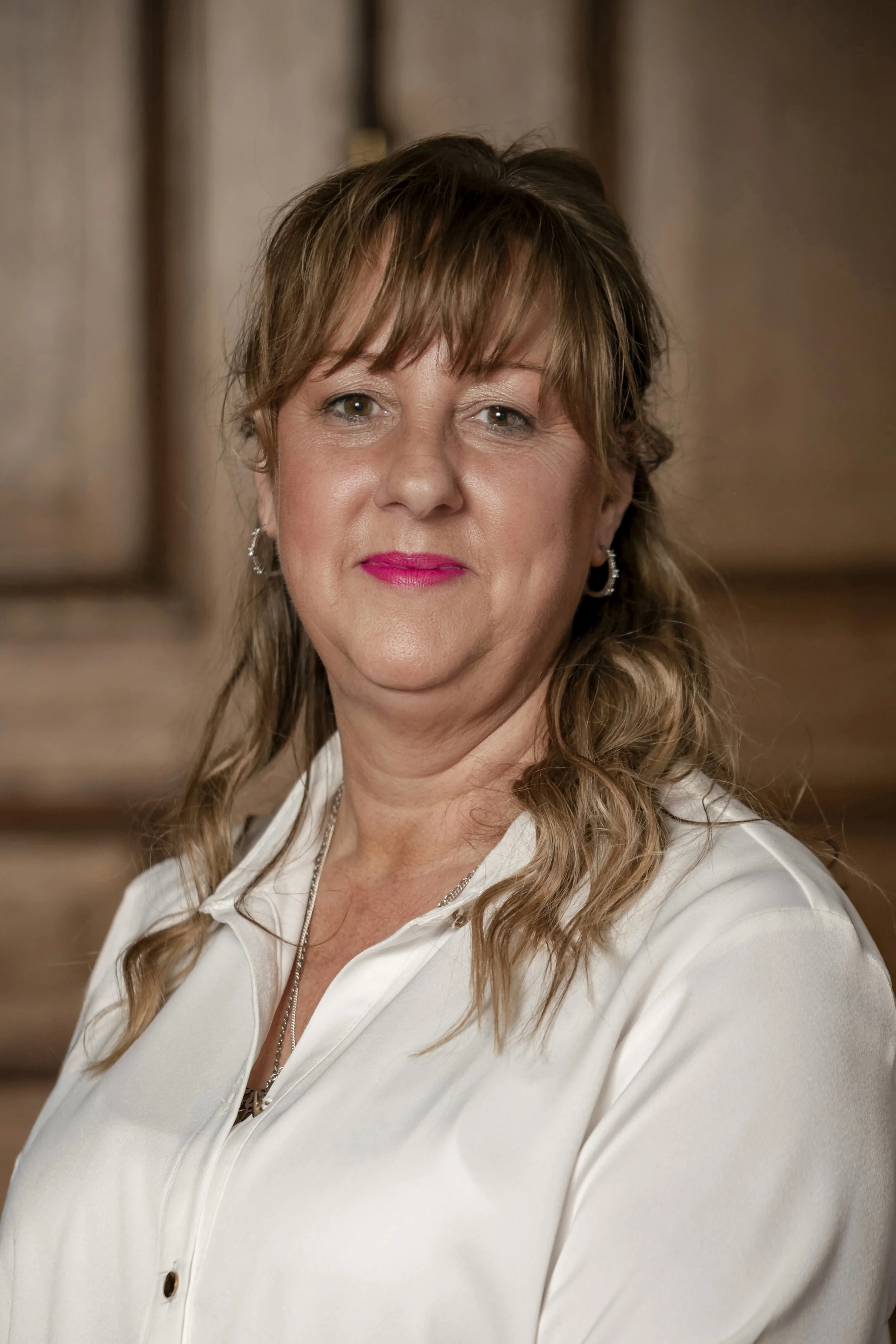 Portrait of a middle-aged woman with light brown wavy hair, wearing a white blouse and pink lipstick, standing in front of a wooden background.