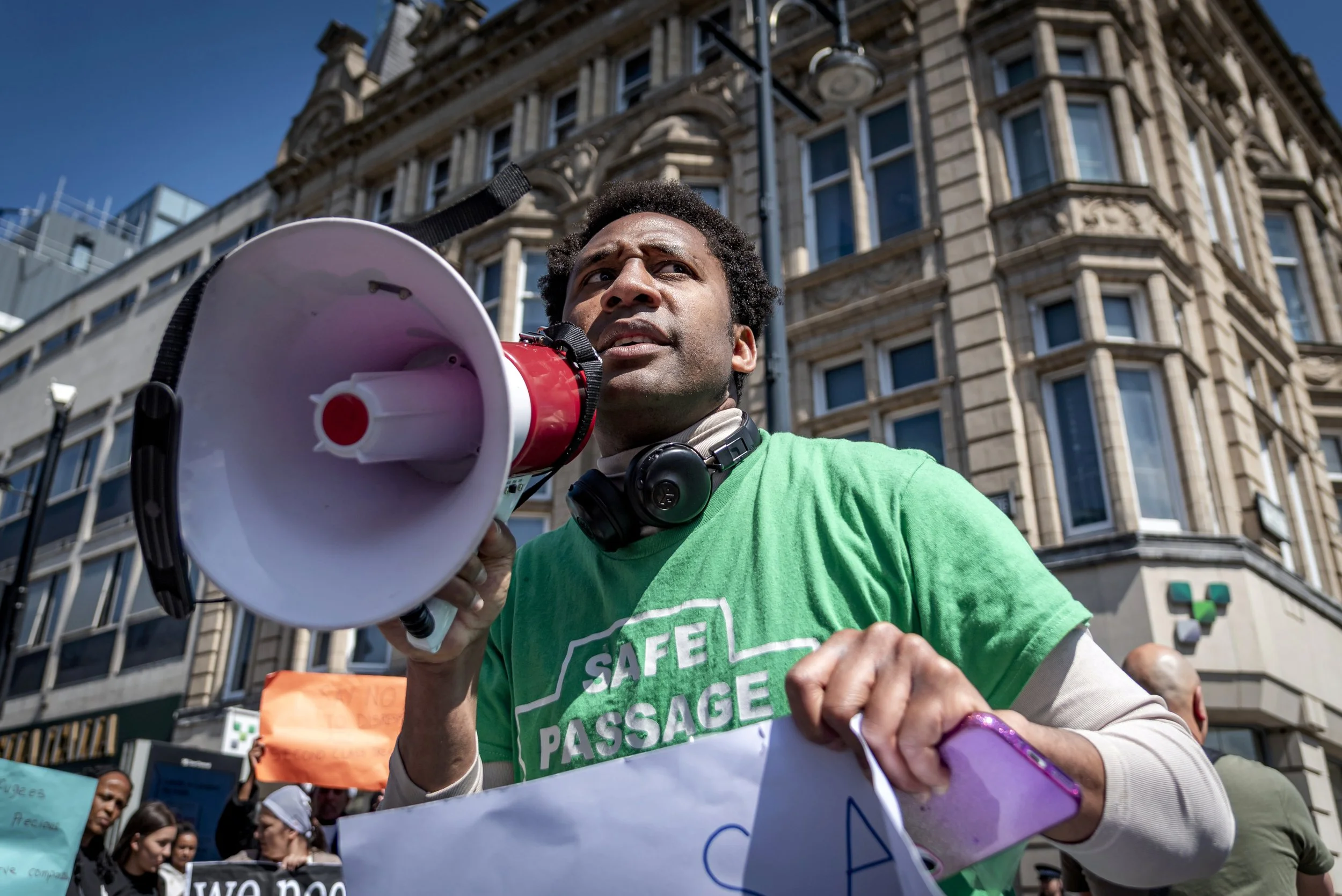 A man wearing a green shirt with the words 'SAFE PASSAGE' on it, holding a megaphone, participating in a protest with other people holding signs, in front of a large historic building.