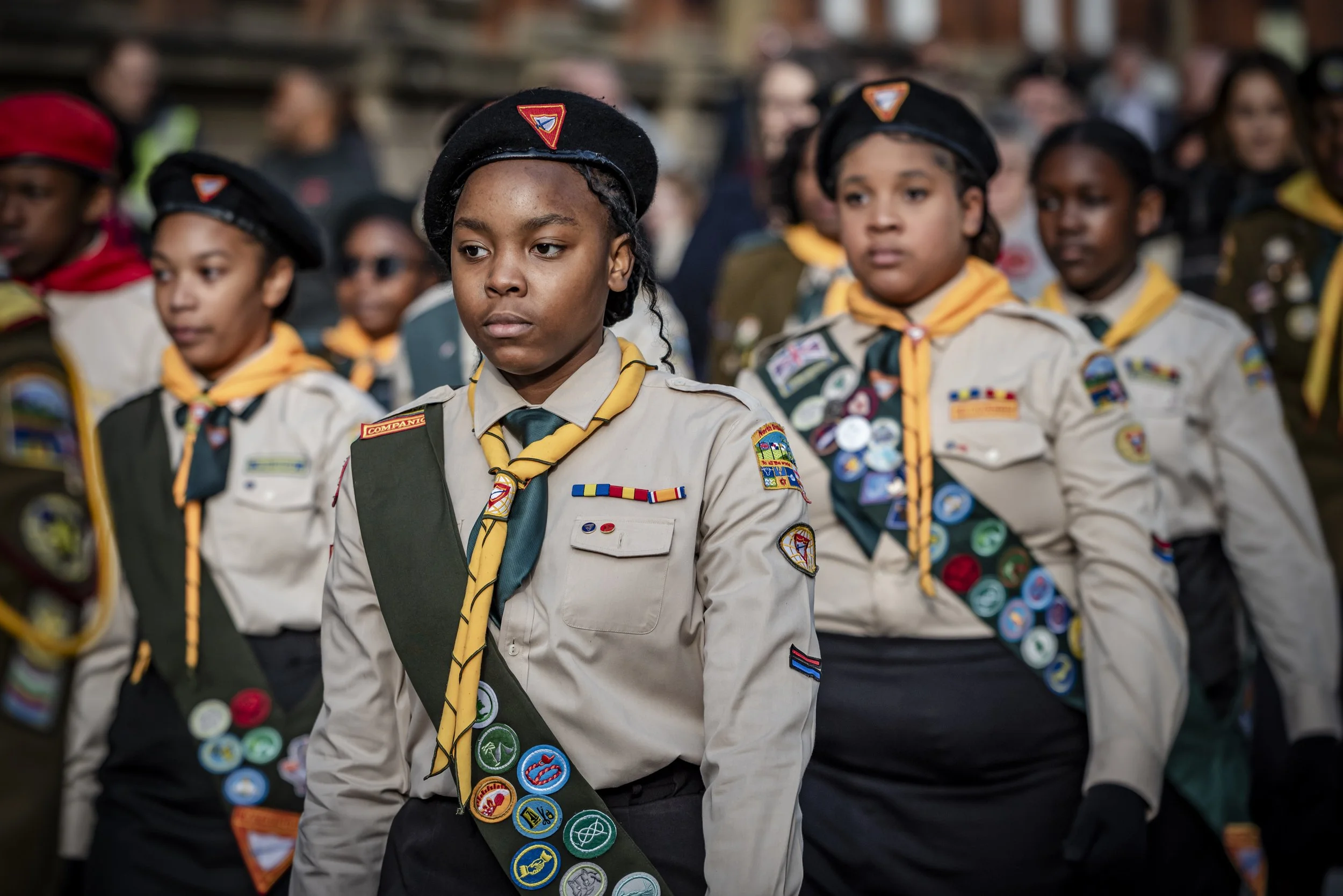 A group of young Girl Scouts in uniform, standing in a formation. They are wearing badges and neckerchiefs, with some also wearing berets. The focus is on one girl at the front with a serious expression, while others are visible in the background.