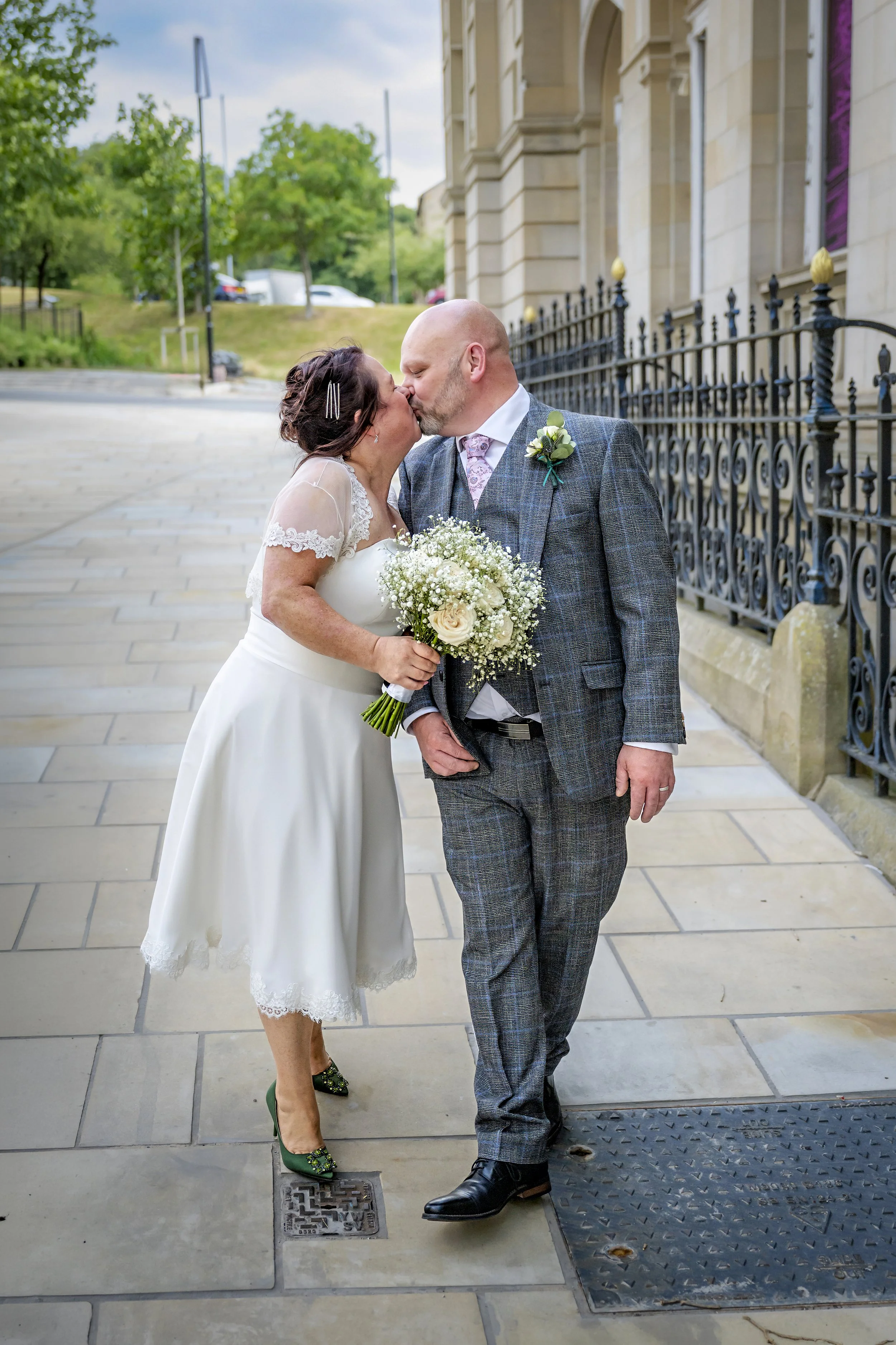 A bride and groom share a kiss, with the bride holding a bouquet of white flowers, outside in front of a historic building with decorative black iron fencing.