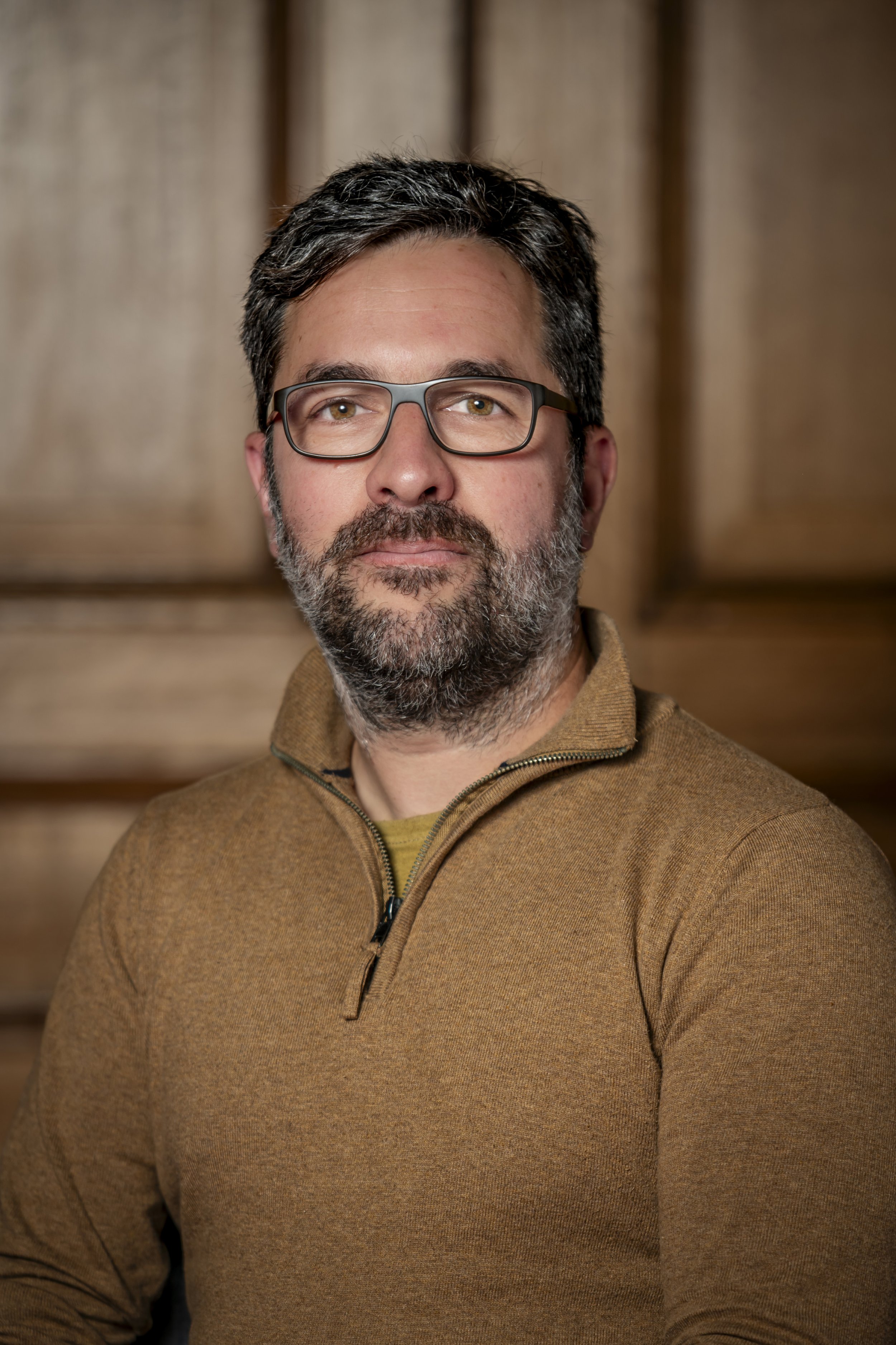A man with glasses and a beard, wearing a brown zip-up sweater, sitting indoors with a wooden background.