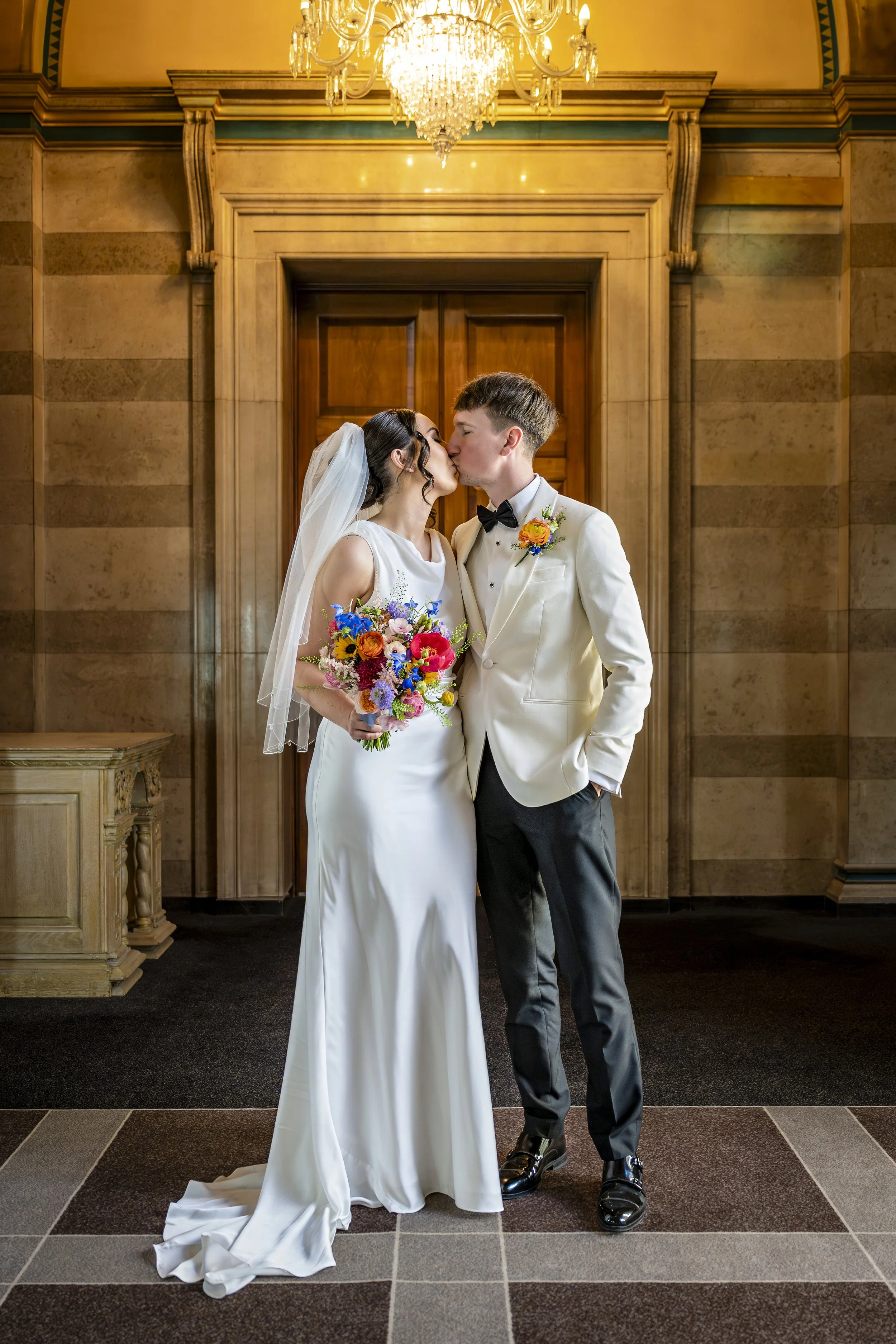 A bride and groom sharing a kiss during their wedding ceremony inside a grand, historic building with a large chandelier and ornate woodwork behind them. The bride holds a colorful bouquet, and the groom is dressed in a white tuxedo jacket with a bou