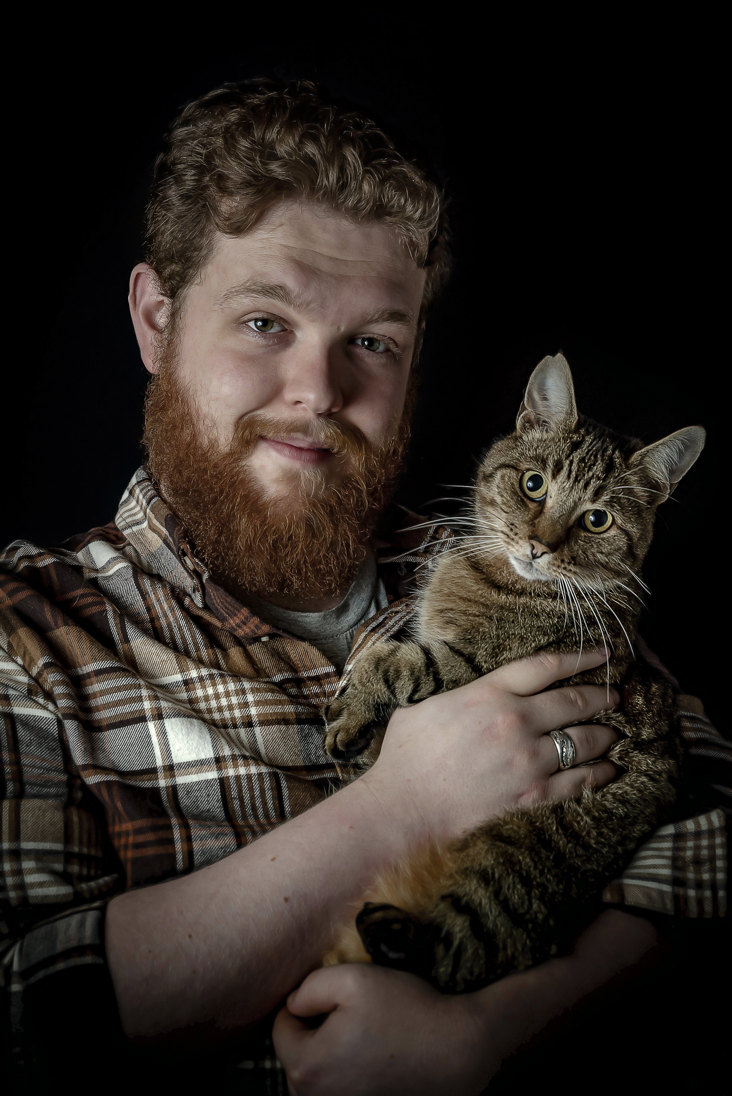 A man with a beard holding a tabby cat against a black background.