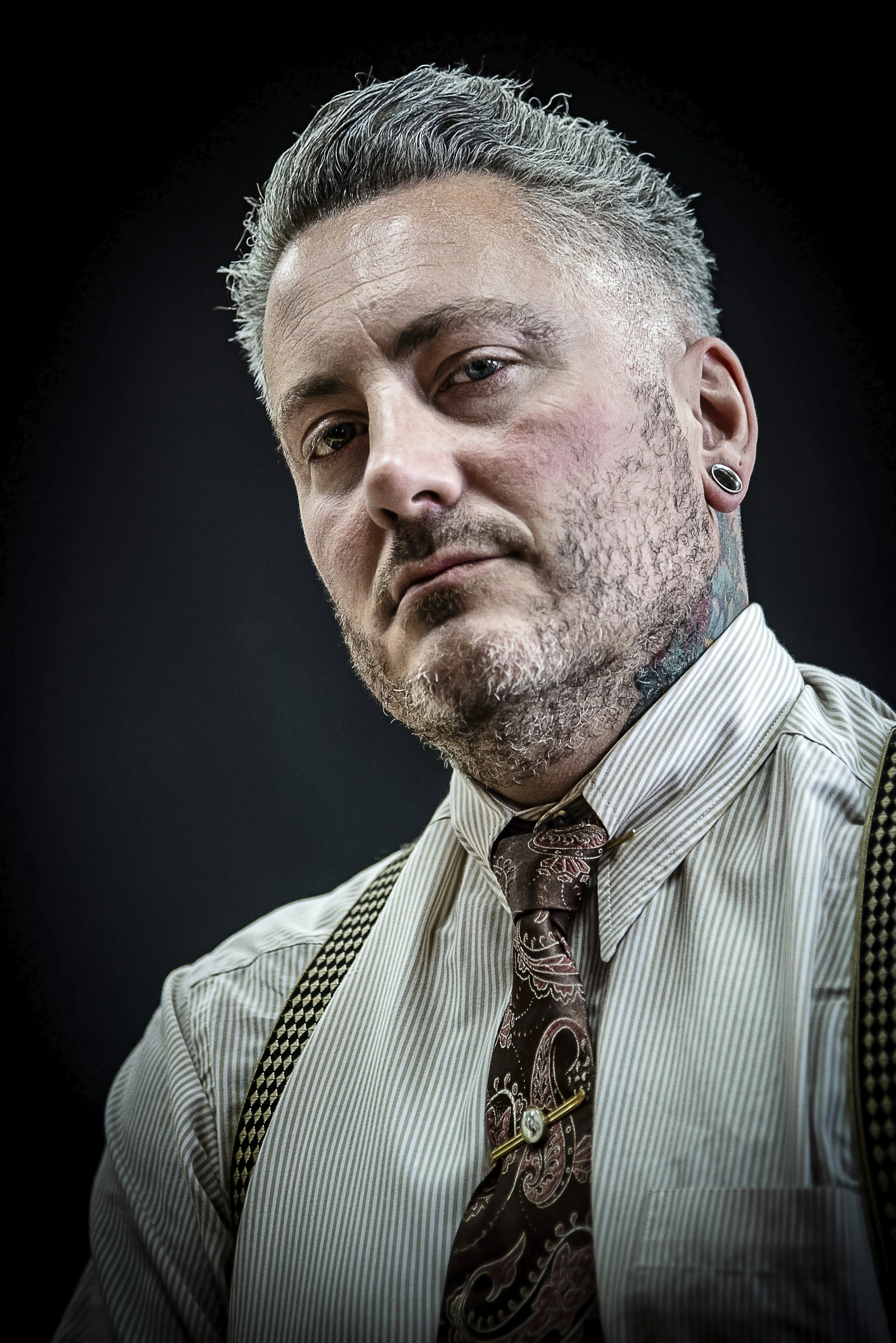 A close-up portrait of a man with styled gray hair, facial stubble, and tattoos on his neck, wearing a striped shirt, patterned tie, suspenders, and a black earring, against a dark background.