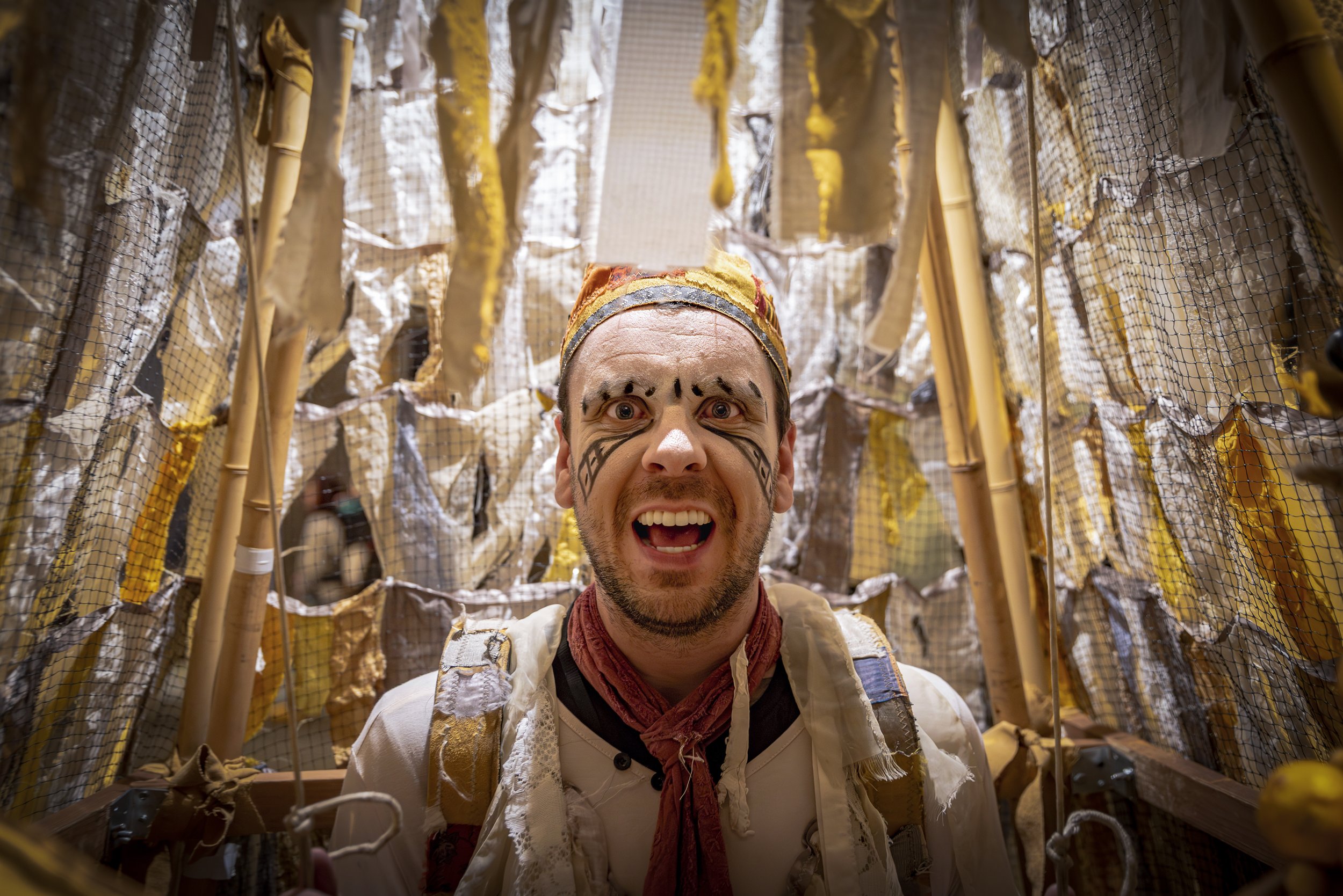 A man dressed as a fictional character with face paint and a colorful headband, standing inside a tunnel made of paper, fabric, and sticks, creating an artistic or theatrical setting.