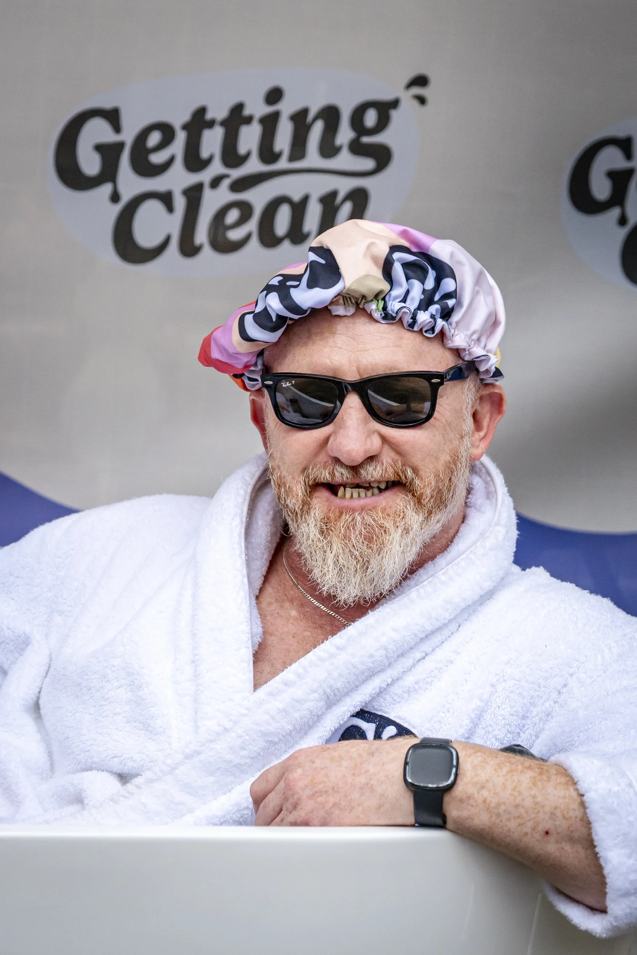 A man smiling while wearing sunglasses, a colorful shower cap, a white towel, and a wristwatch, sitting in front of a sign that says 'Getting Clean'.