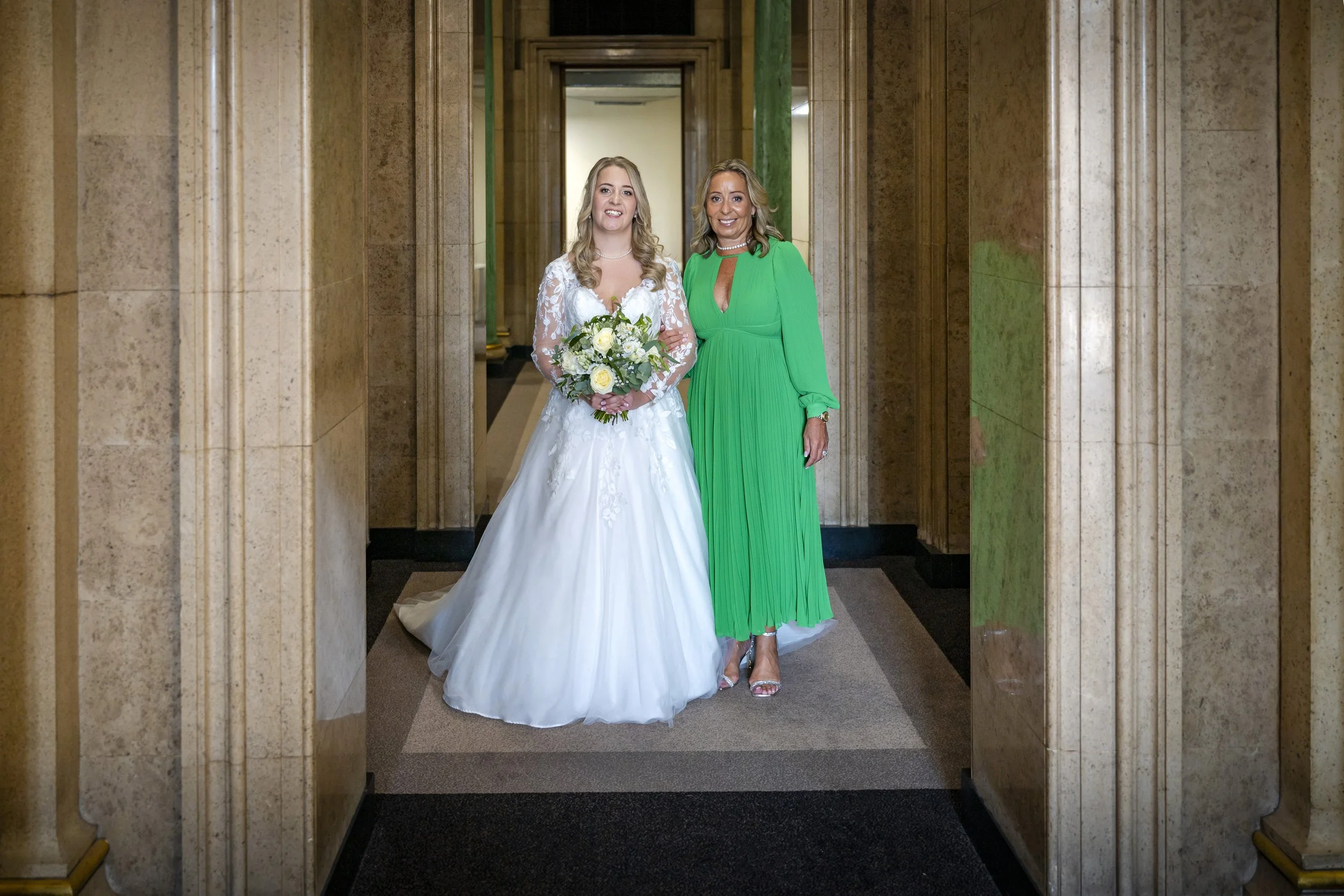 A bride in a white wedding gown holding a bouquet standing next to a woman in a bright green dress inside a building with marble walls.
