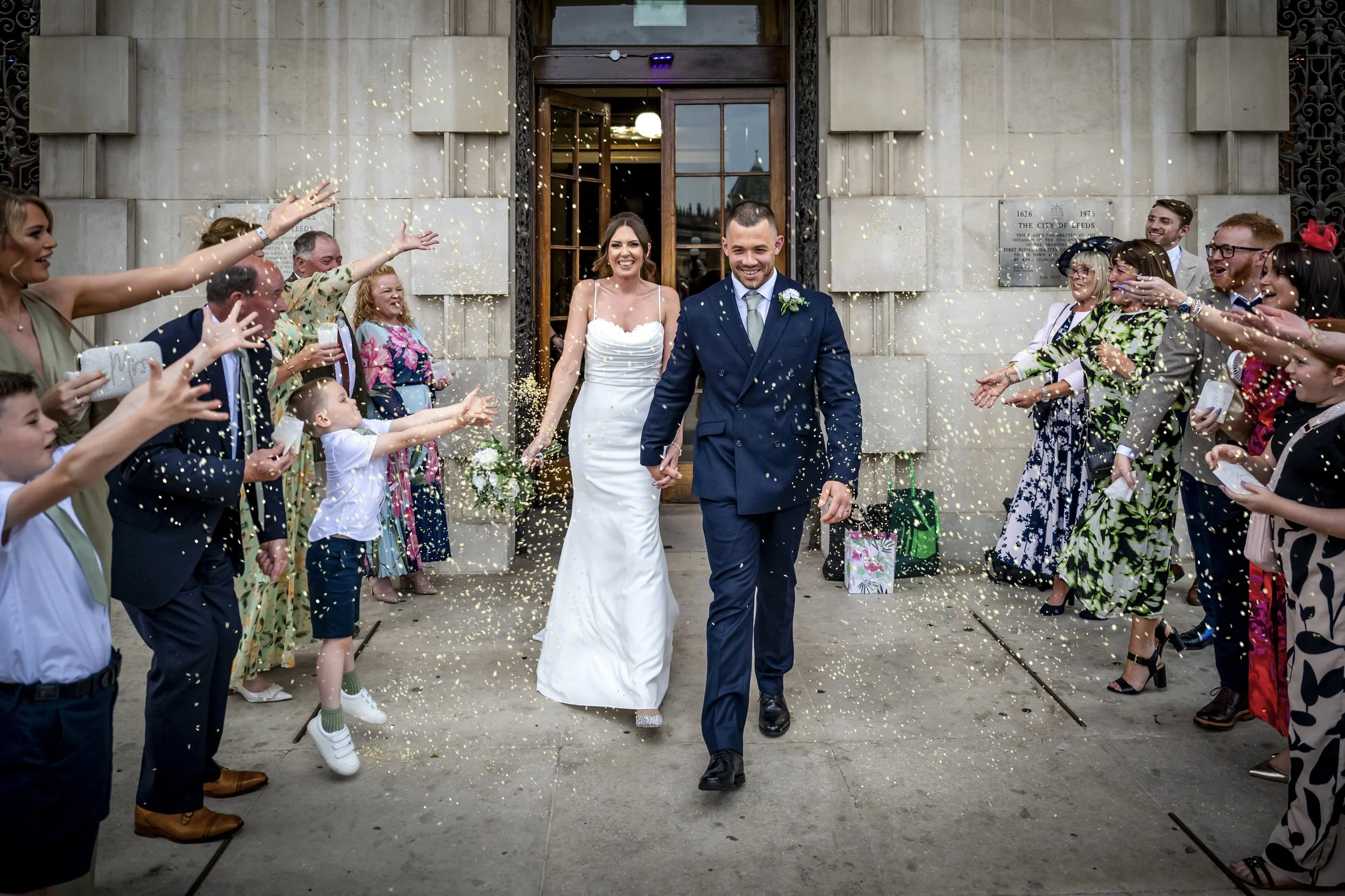A newlywed couple walking out of a building, smiling, as guests throw confetti and cheer.