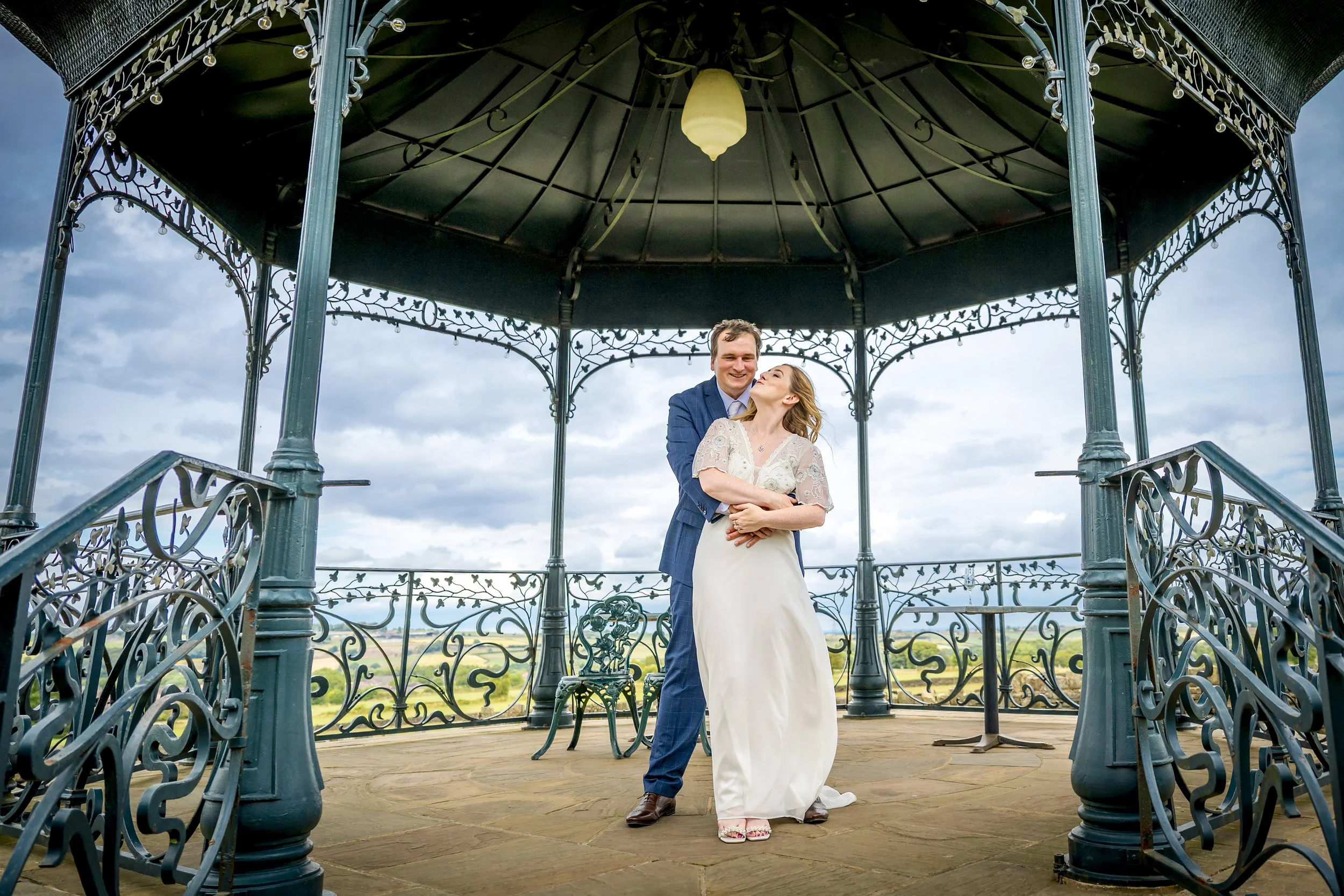 A bride and groom standing underneath a ornate, open-air, metal gazebo on a cloudy day, embracing each other.