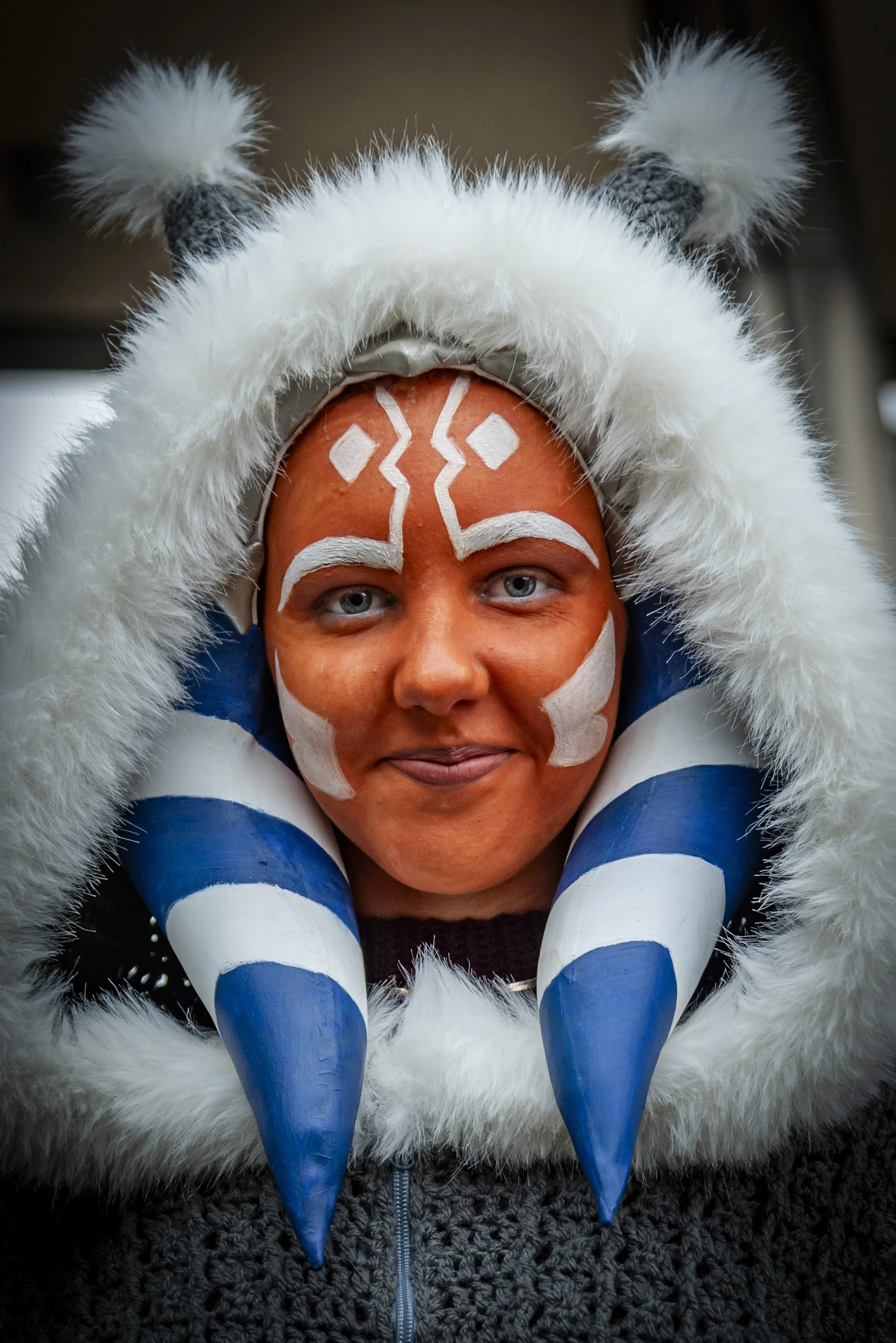 Person wearing a costume with face paint and a headpiece resembling a creature with blue and white striped horns and white fur, in front of a plain background.