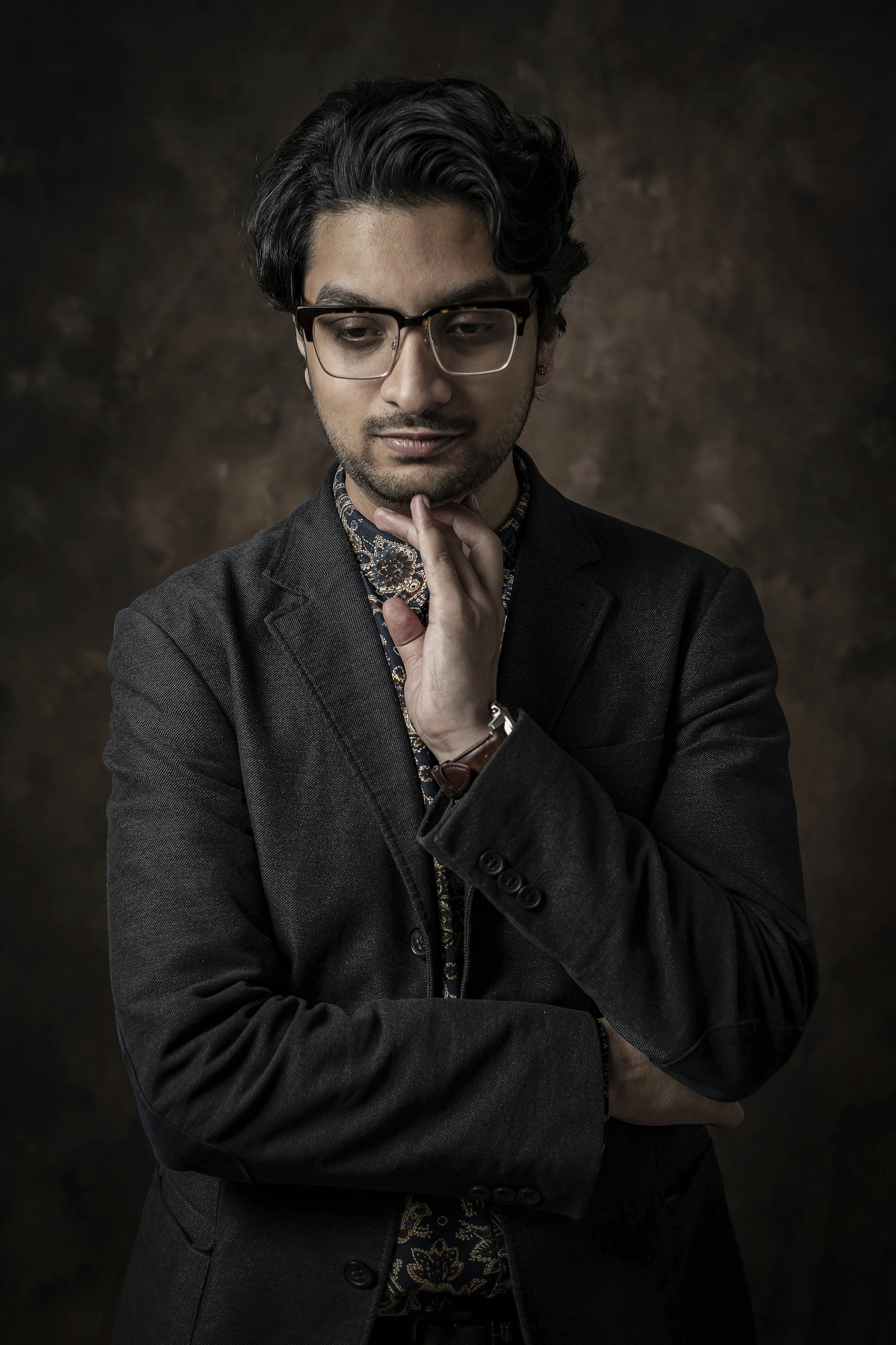 A man with glasses, dark wavy hair, and light skin wearing a dark blazer over a patterned shirt, touching his chin with his hand, standing against a brown textured background.
