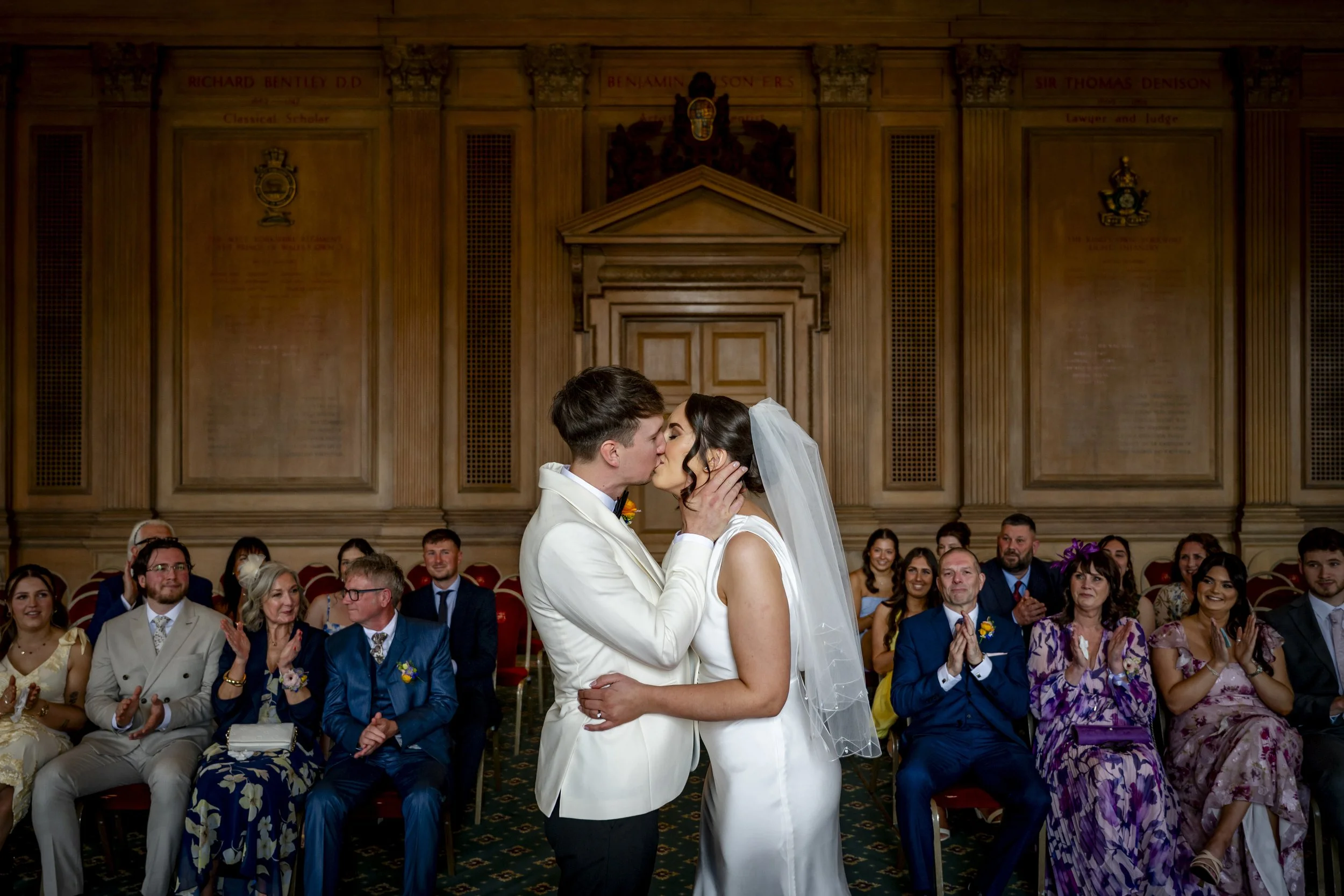 A bride and groom kiss during their wedding ceremony in a grand room with wood-paneled walls and a large audience of diverse guests applauding.